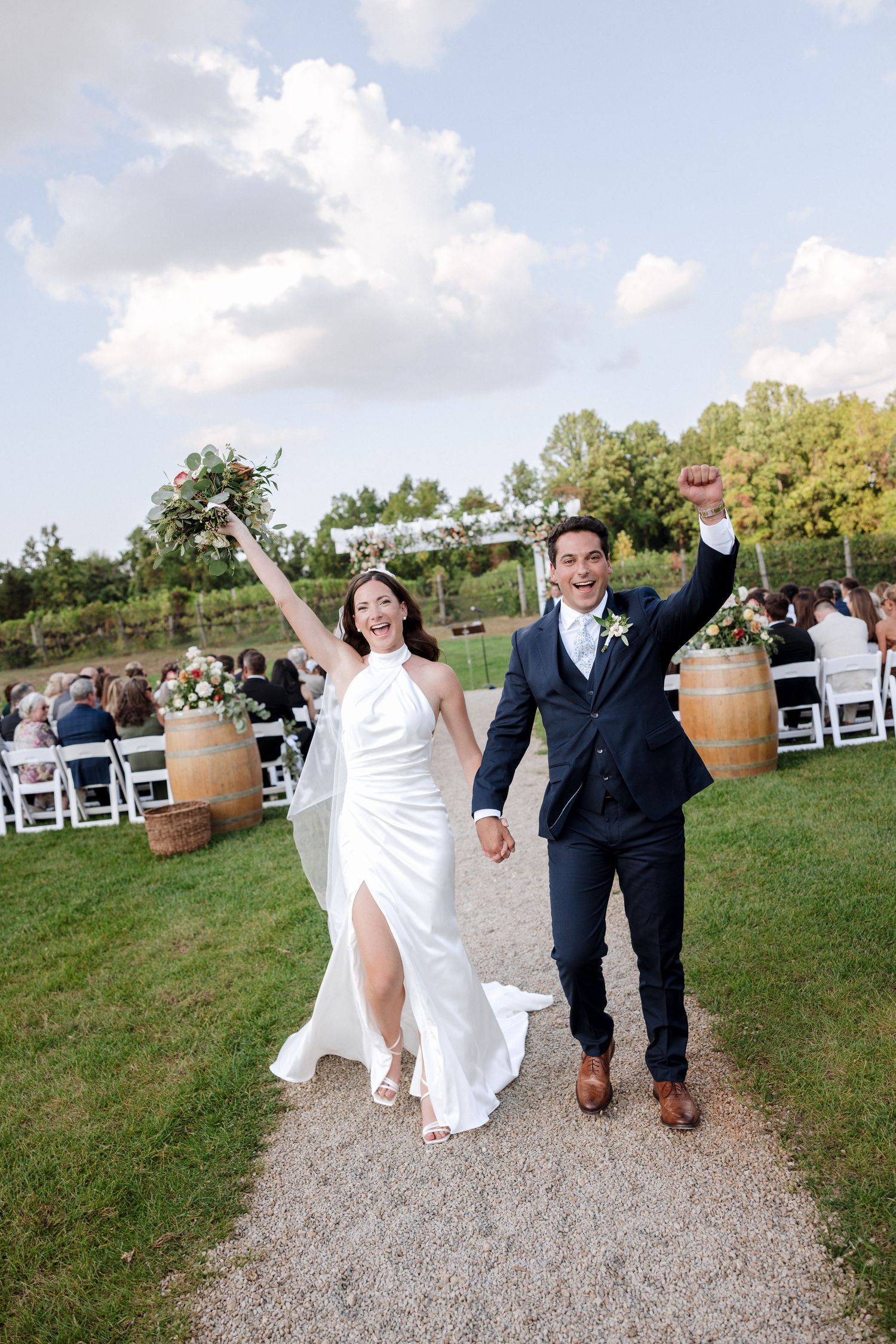 Joyful newlyweds celebrating recessional on gravel path at outdoor vineyard wedding, arms raised in triumph.