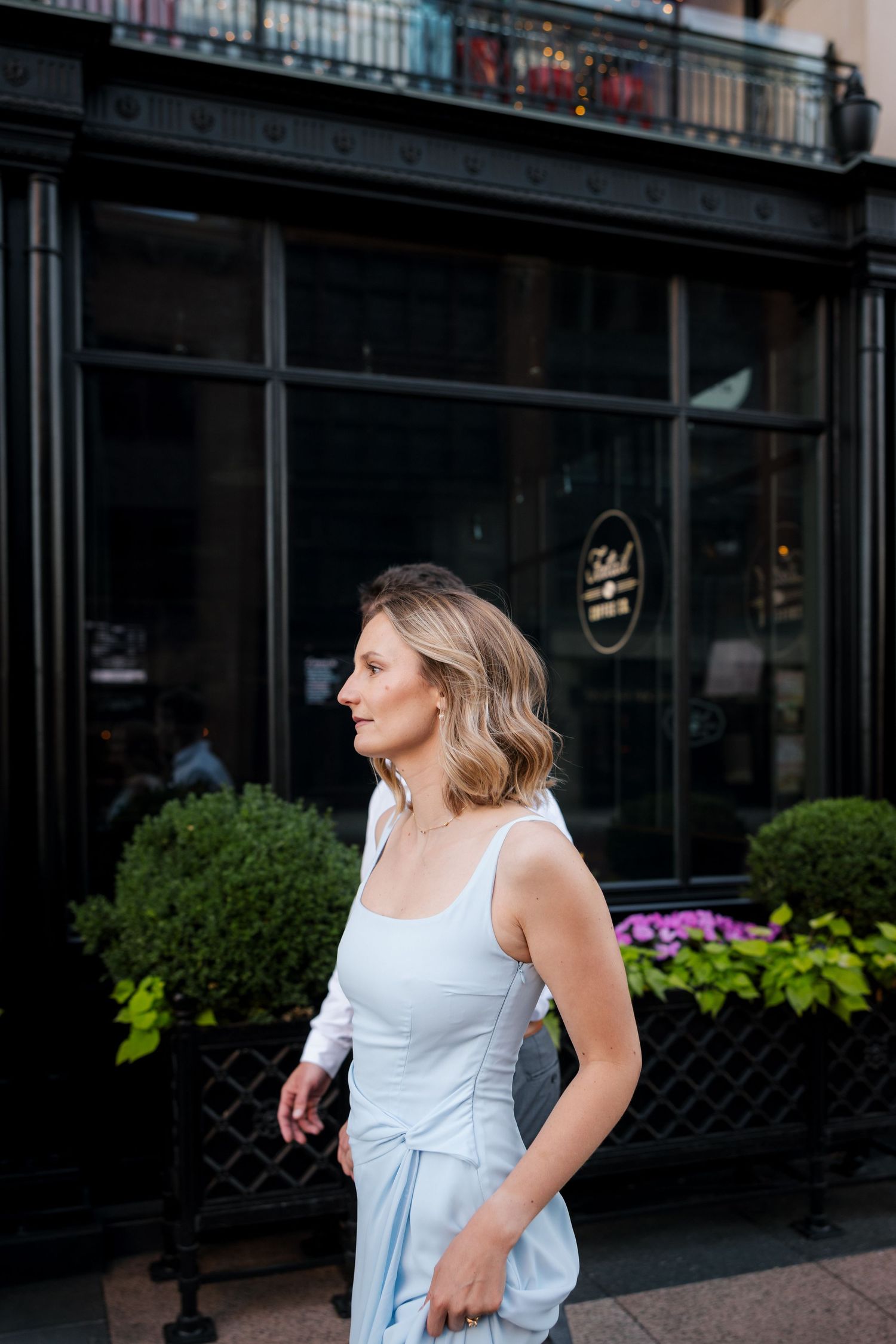 Elegant woman in light blue sleeveless dress standing outside a classic black-framed storefront.
