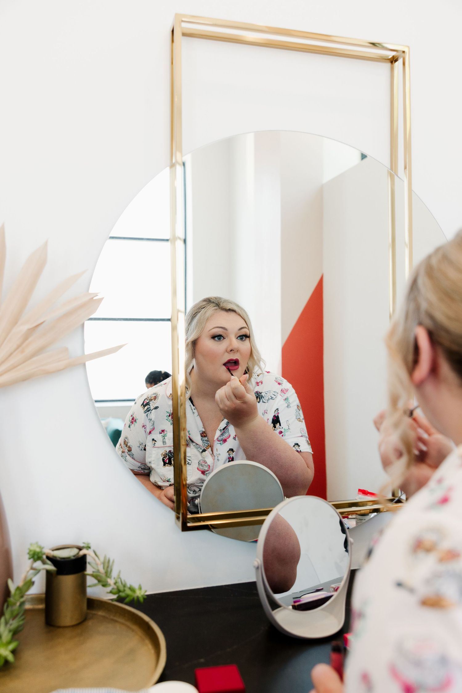 Blonde woman applying red lipstick while looking in a round mirror on a vanity table.