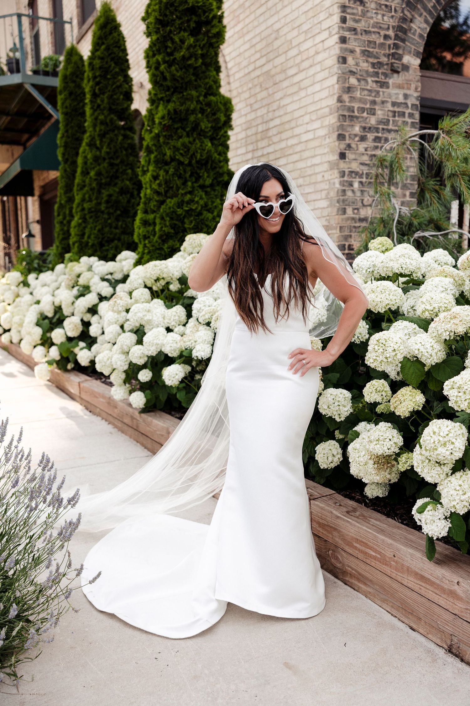 Bride in white mermaid gown and veil poses with heart sunglasses among white hydrangeas outdoors.