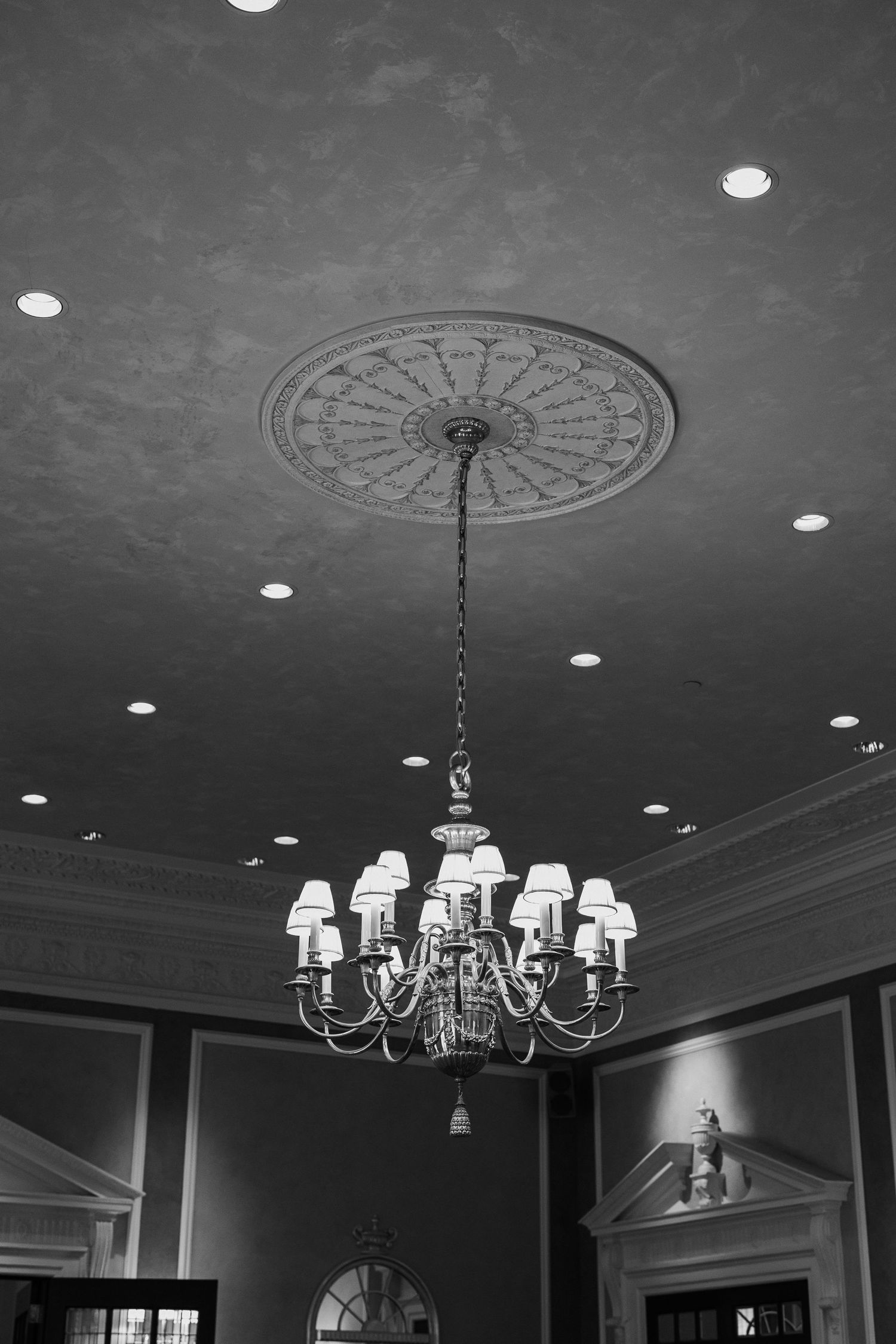 Black and white photo of an ornate chandelier hanging from a decorative ceiling medallion in an elegant venue.