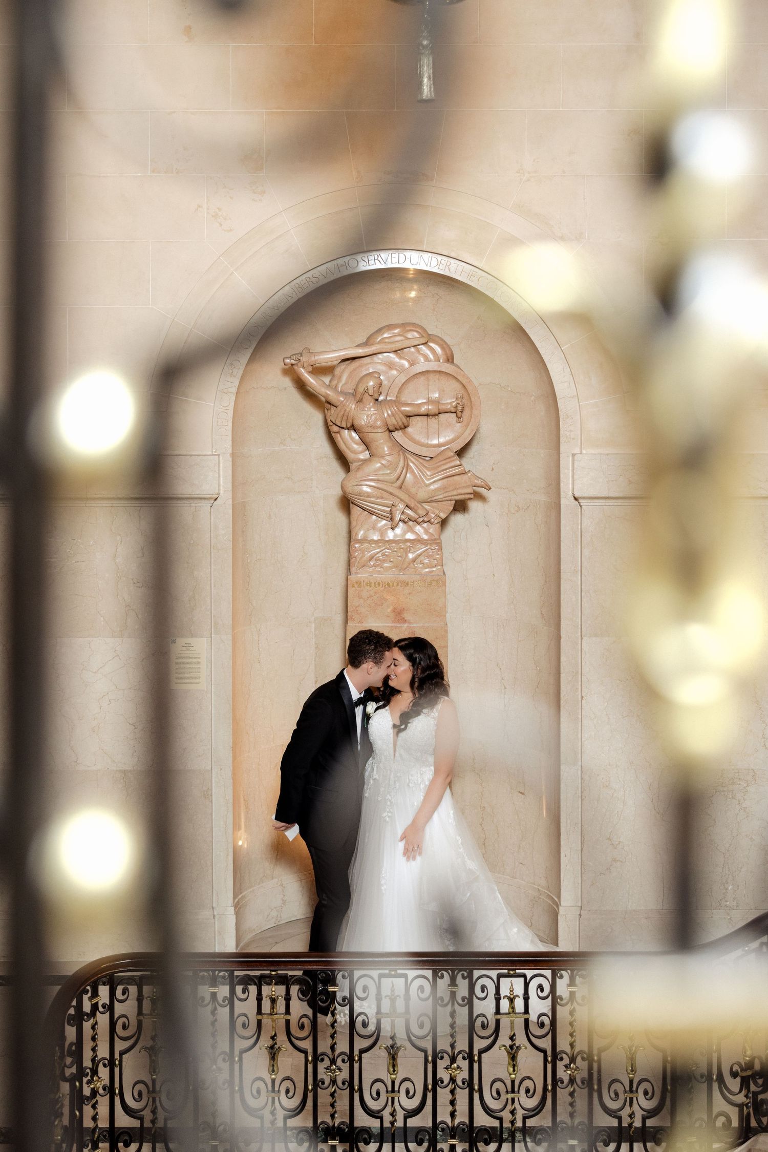Bride and groom kissing beneath a decorative stone warrior sculpture, framed by bokeh lights and iron railing.