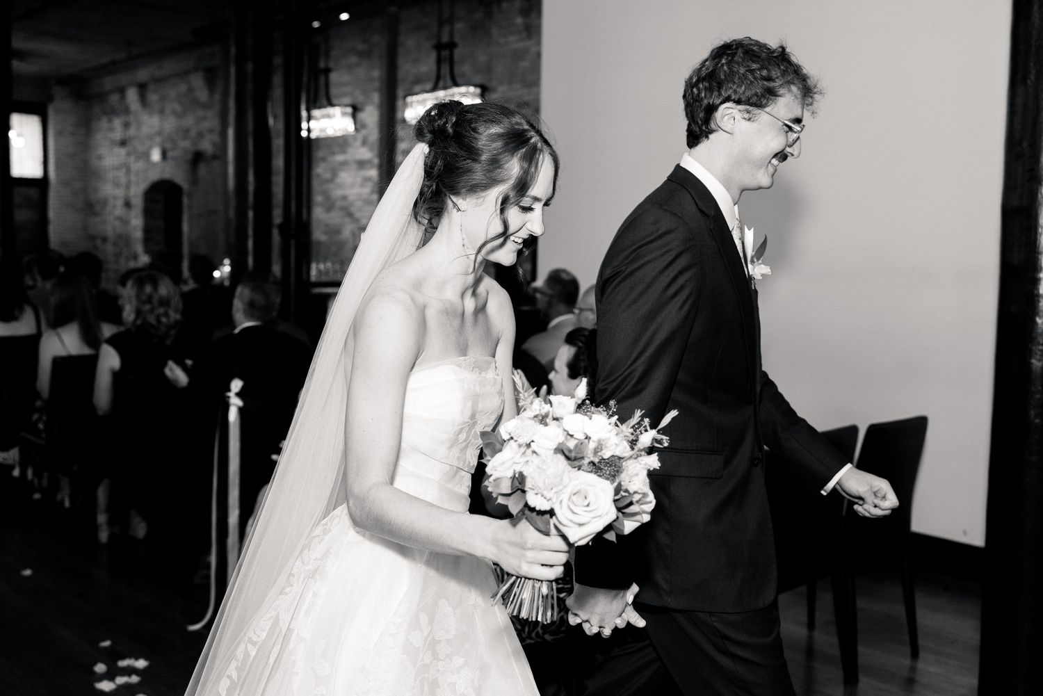 Black and white photo of a smiling bride in veil walking down the aisle holding a white bouquet.