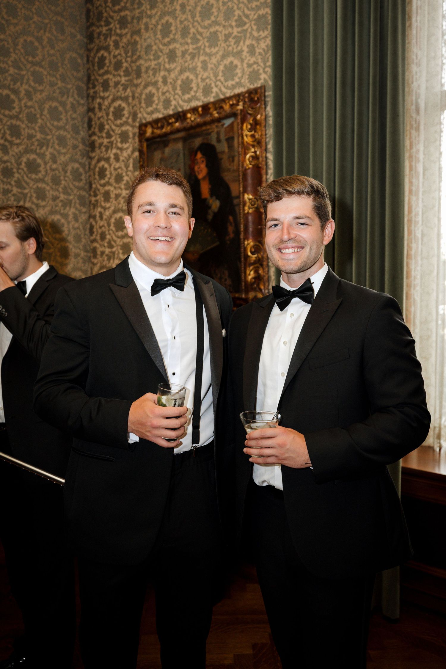 Two smiling men in black tuxedos holding cocktails at a formal black tie wedding reception.