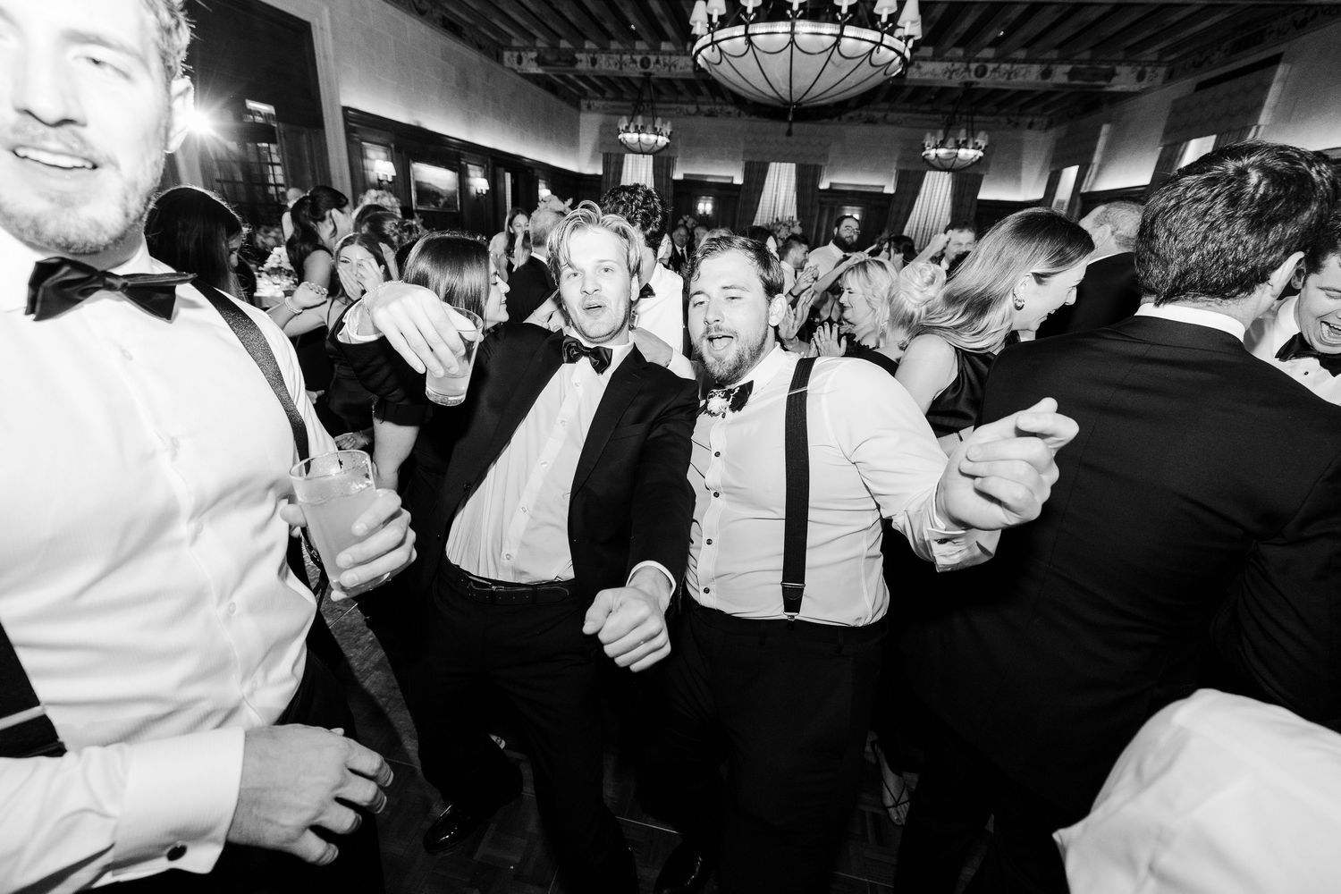 Black and white photo of men in tuxedos dancing joyfully at a formal wedding reception party.