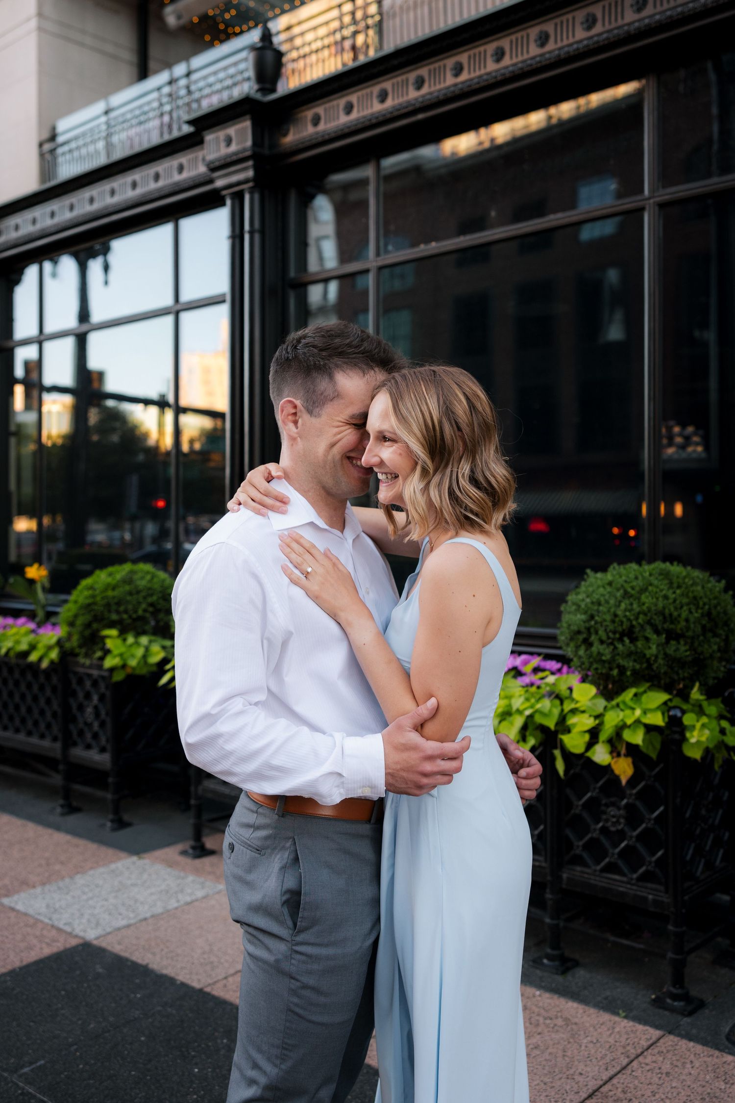 Happy couple embracing and smiling outside a dark-windowed building with green flower boxes.