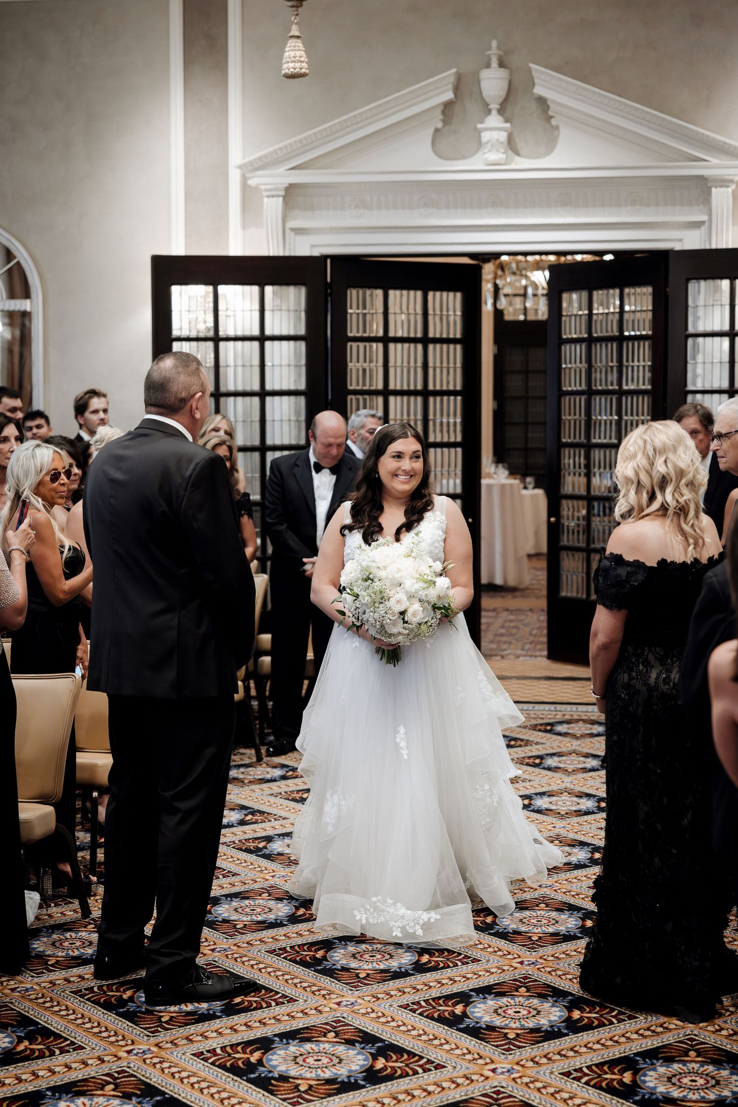 Bride walks down the aisle in a white ball gown holding a white floral bouquet during an elegant indoor wedding ceremony.