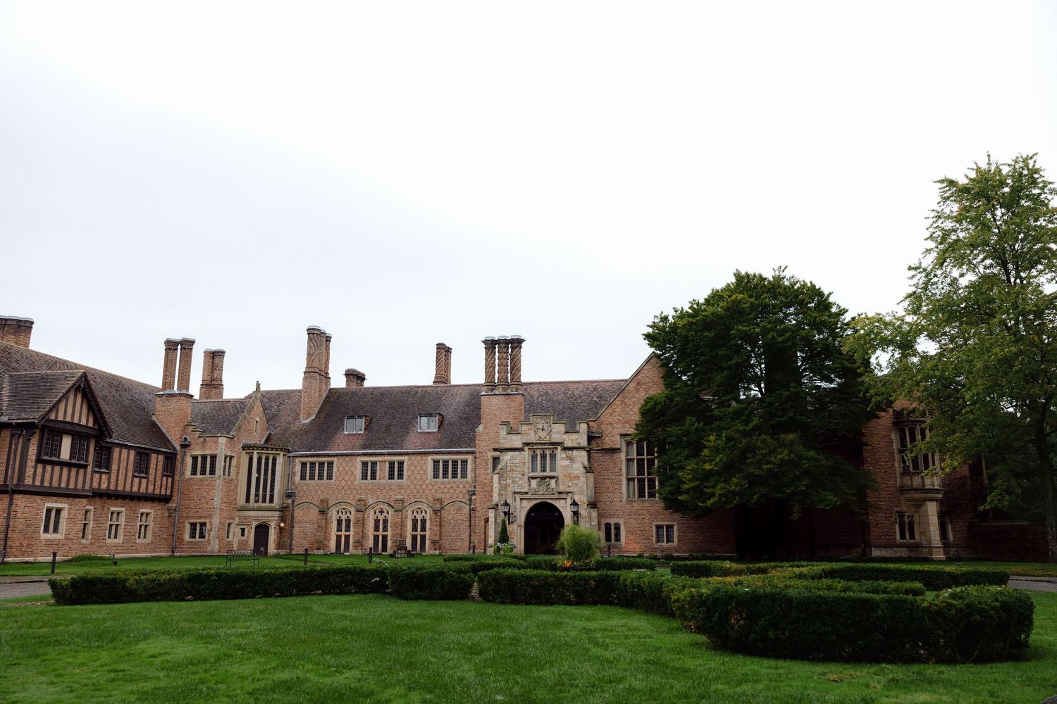 Historic Tudor-style manor house with red brick facade, multiple chimneys, manicured green lawns, and lush trees under overcast sky.