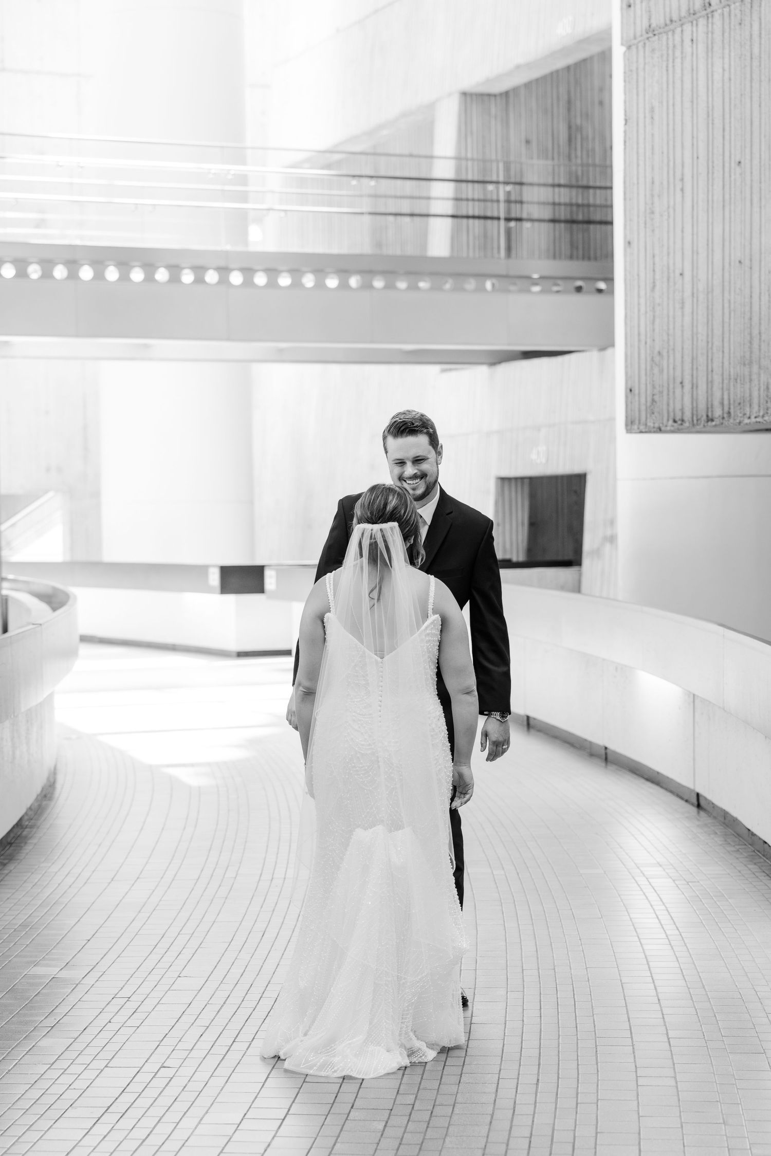 Black and white photo of smiling groom in suit walking with bride in white gown and veil in elegant venue.
