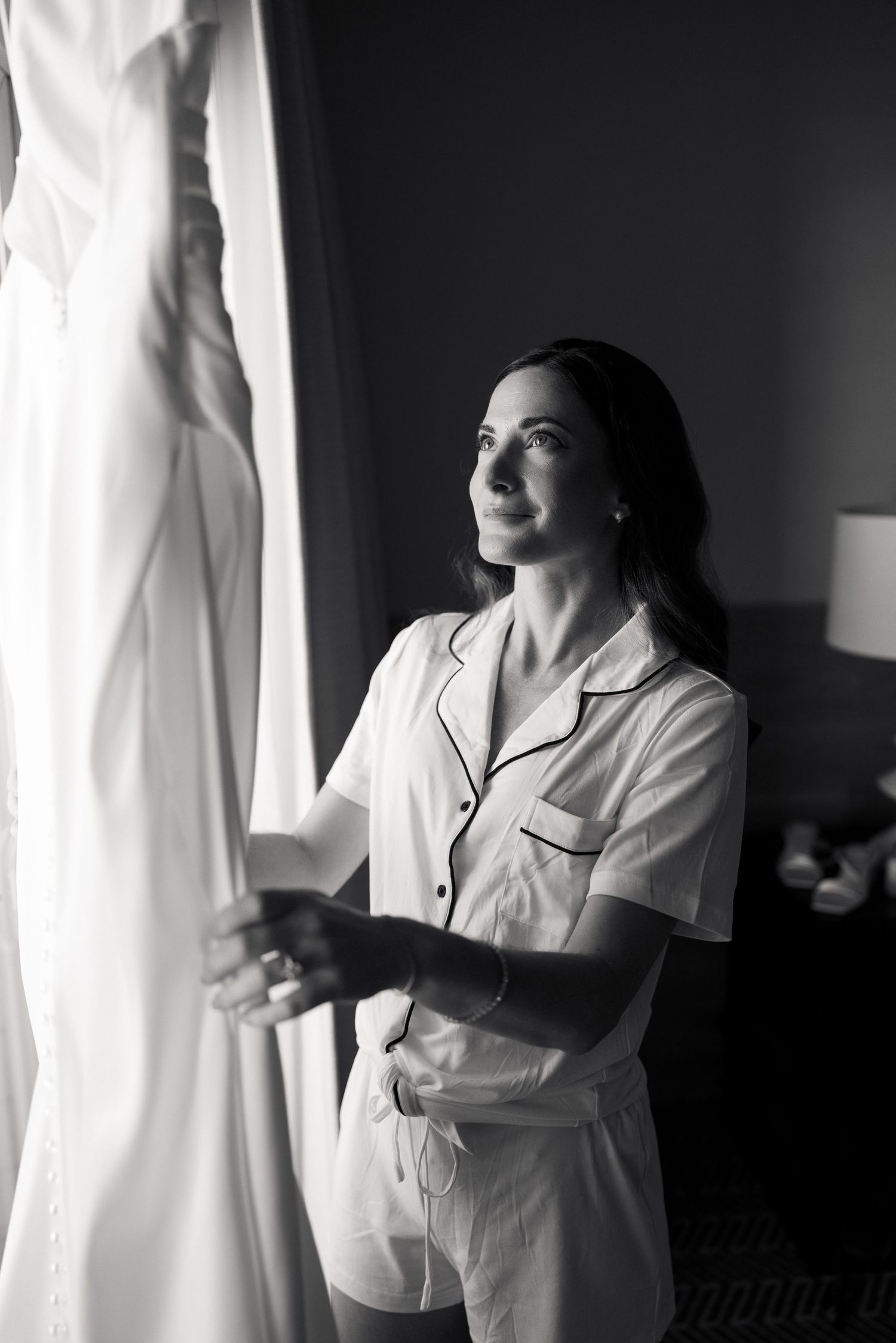 Bride in white pajamas gazing at her wedding dress hanging by a window in black and white photo.