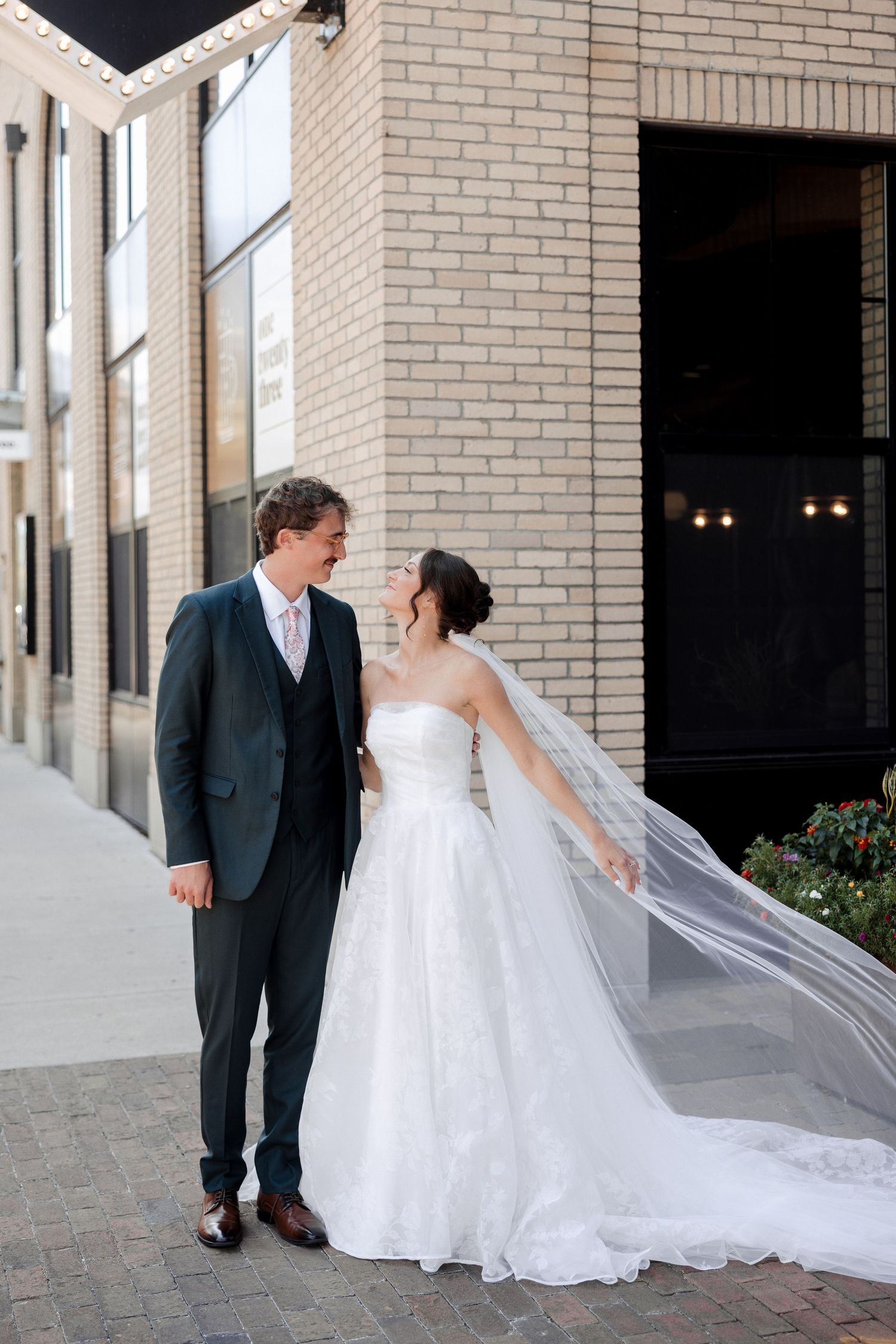 A bride in a flowing white gown and veil stands with a groom in a dark suit, smiling at each other outside a brick building.
