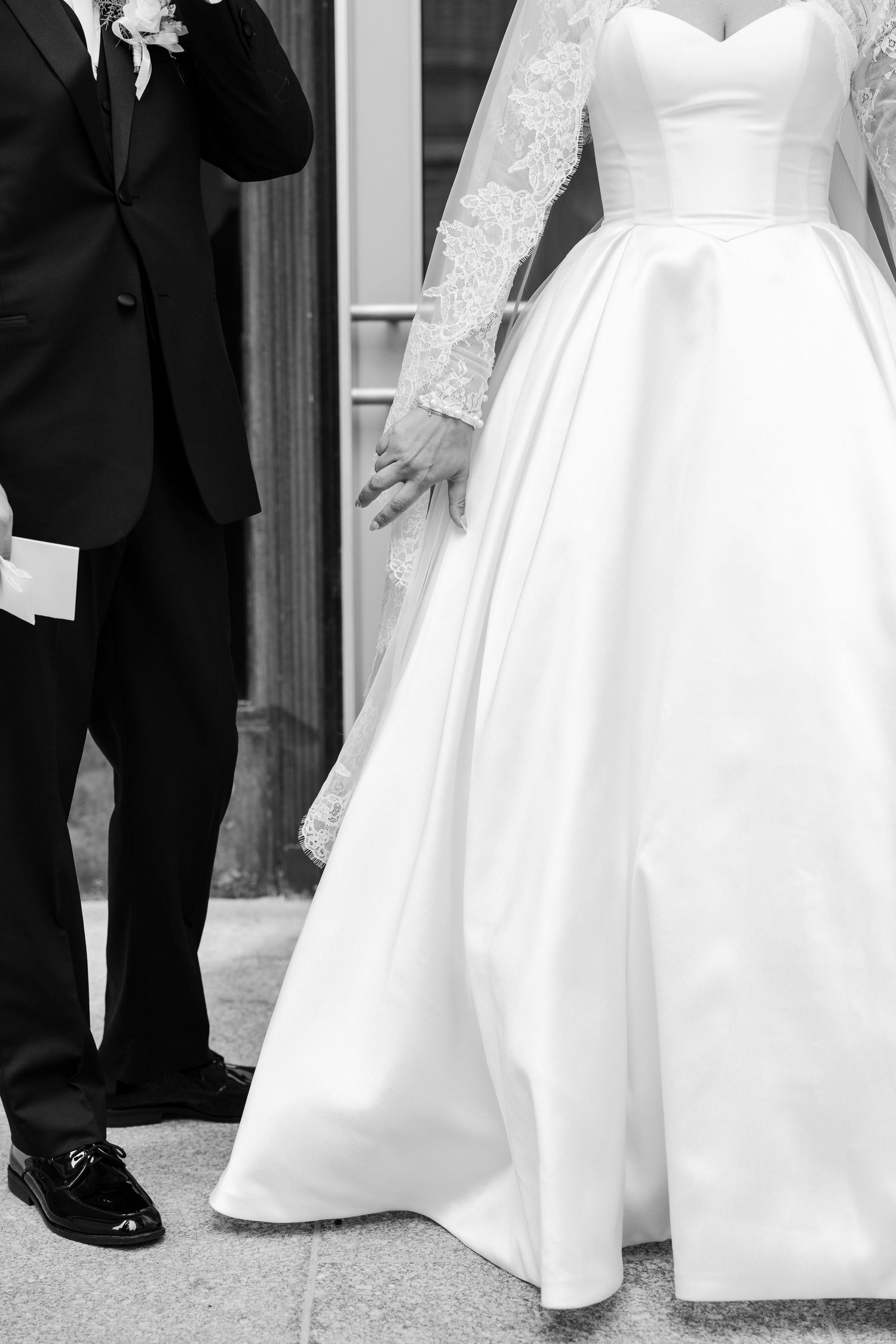 Black and white photo of bride and groom holding hands, bride in lace ball gown, groom in tuxedo.