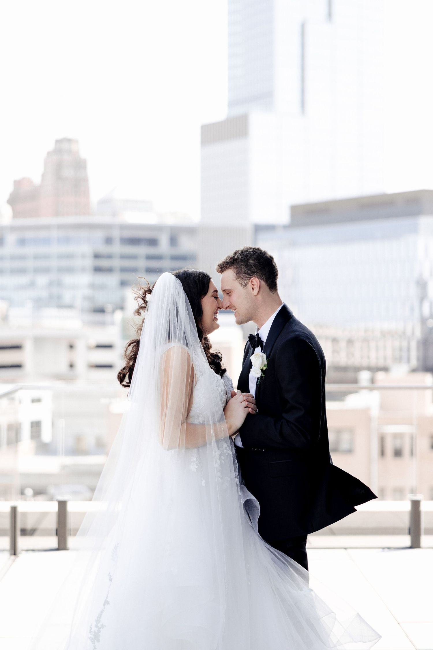 Bride and groom lean in close smiling on a rooftop, surrounded by a city skyline on their wedding day.
