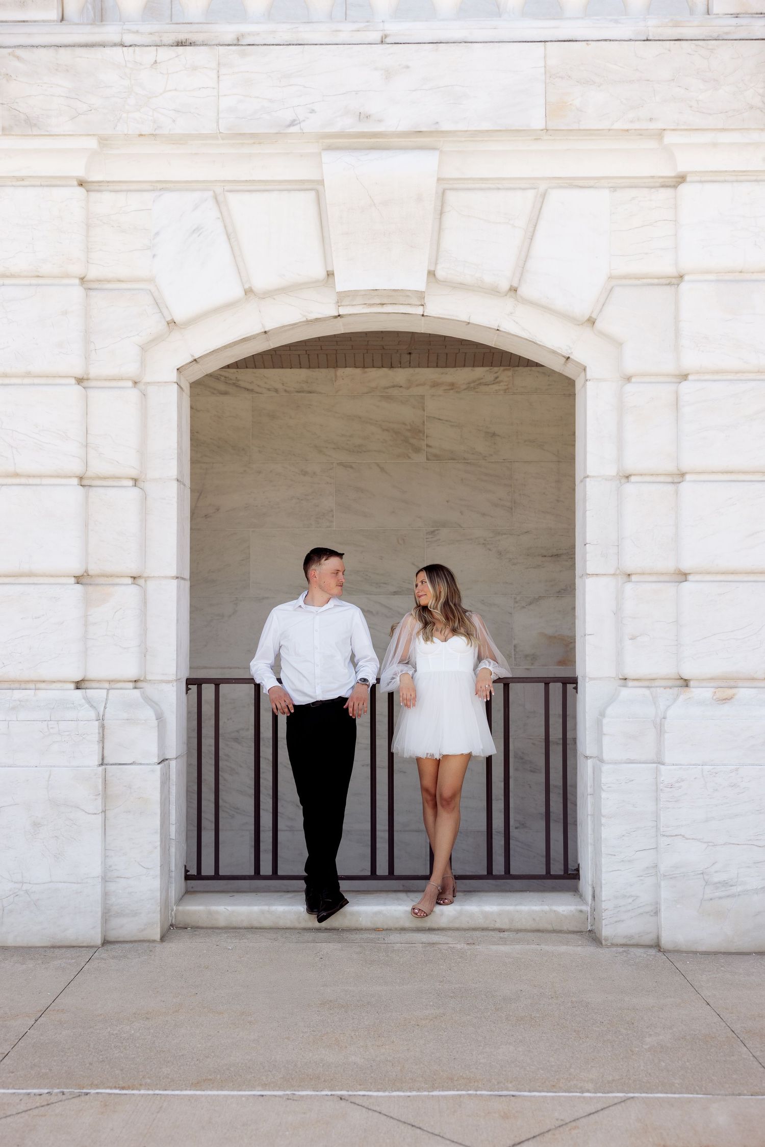 Couple in white outfits holding hands under a marble arch at an elegant building, sharing a romantic glance.