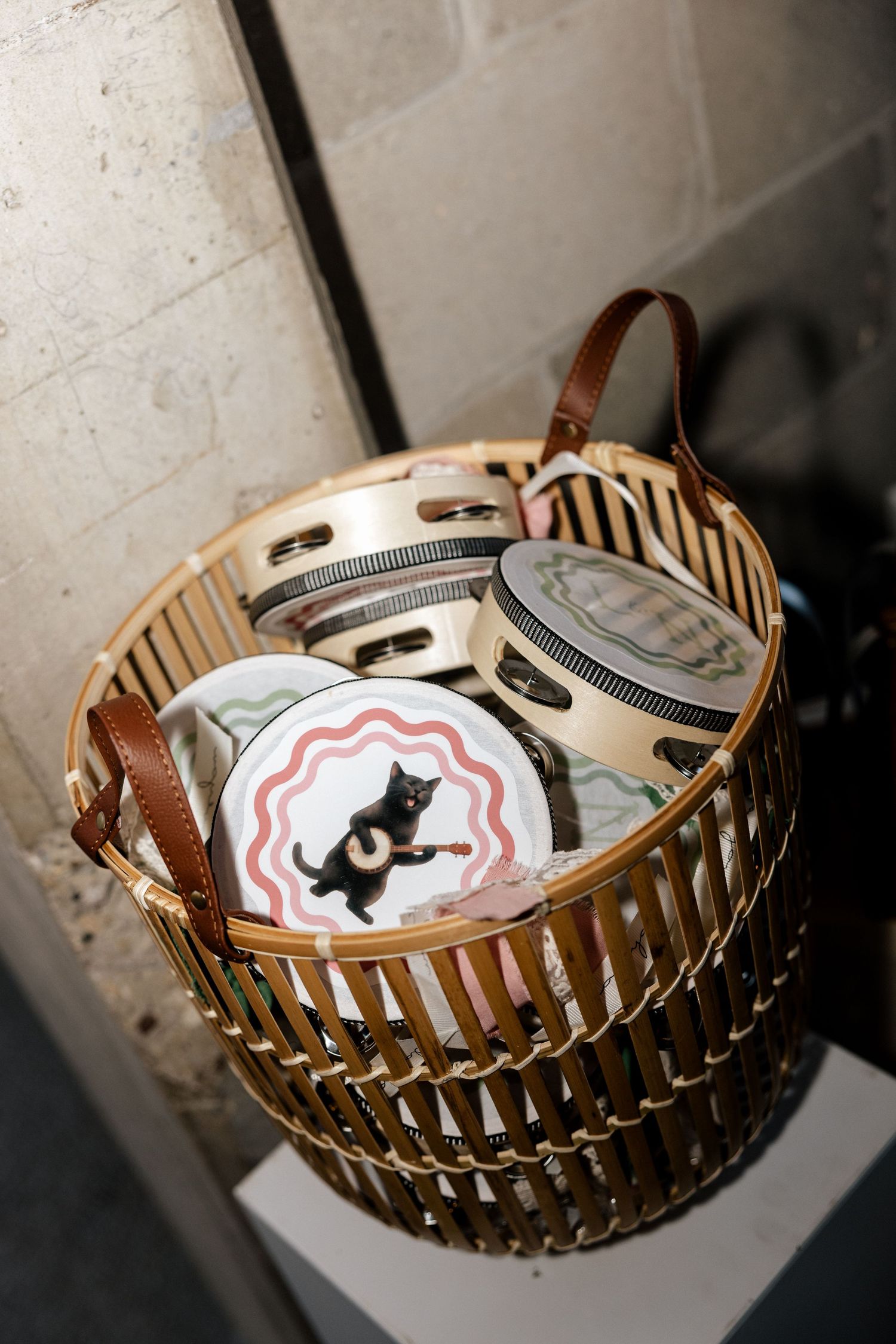 Wicker basket with leather handles holding decorative round tins featuring cat and floral designs.