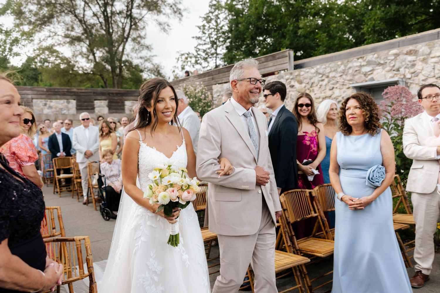 Bride walks down outdoor aisle with escort, holding floral bouquet, guests seated on wooden chairs at sunny wedding ceremony.
