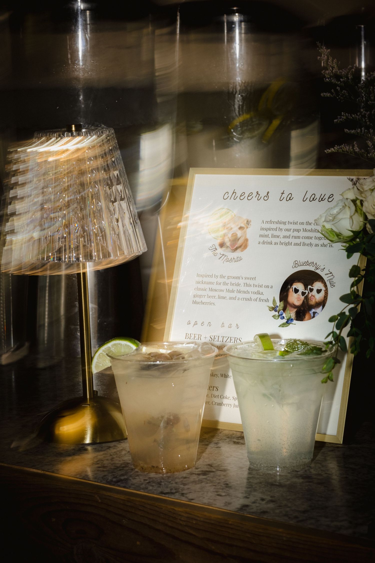 Wedding signature cocktails displayed with a cheers to love sign, lime garnish, and decorative lamp at a reception bar.