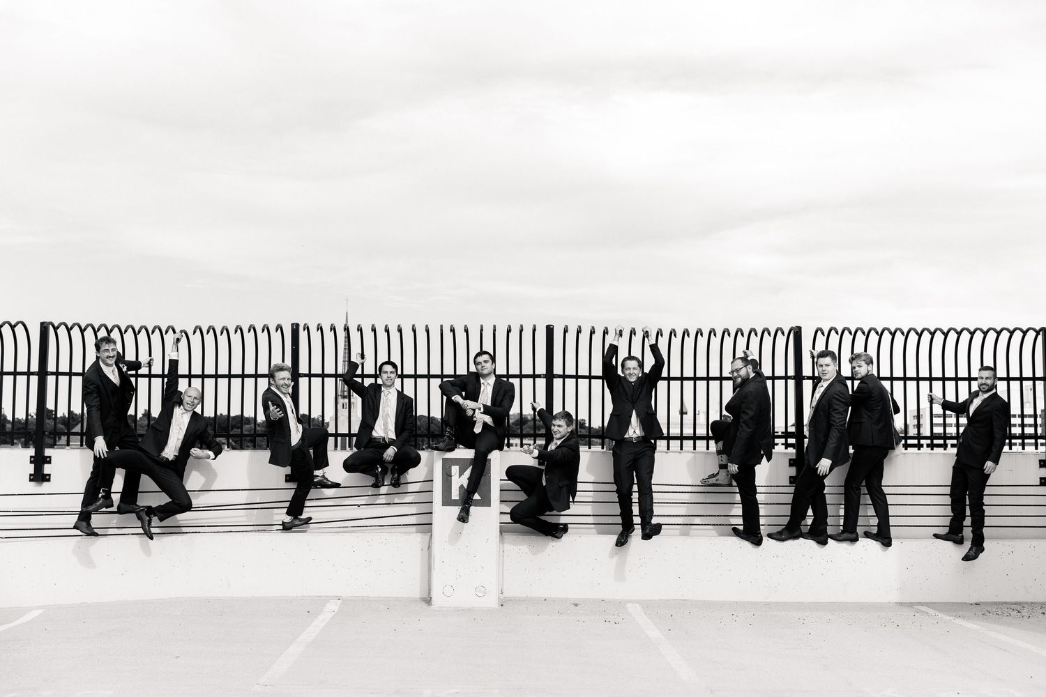 Black and white photo of groomsmen jumping and posing on a rooftop fence in suits at a wedding celebration.