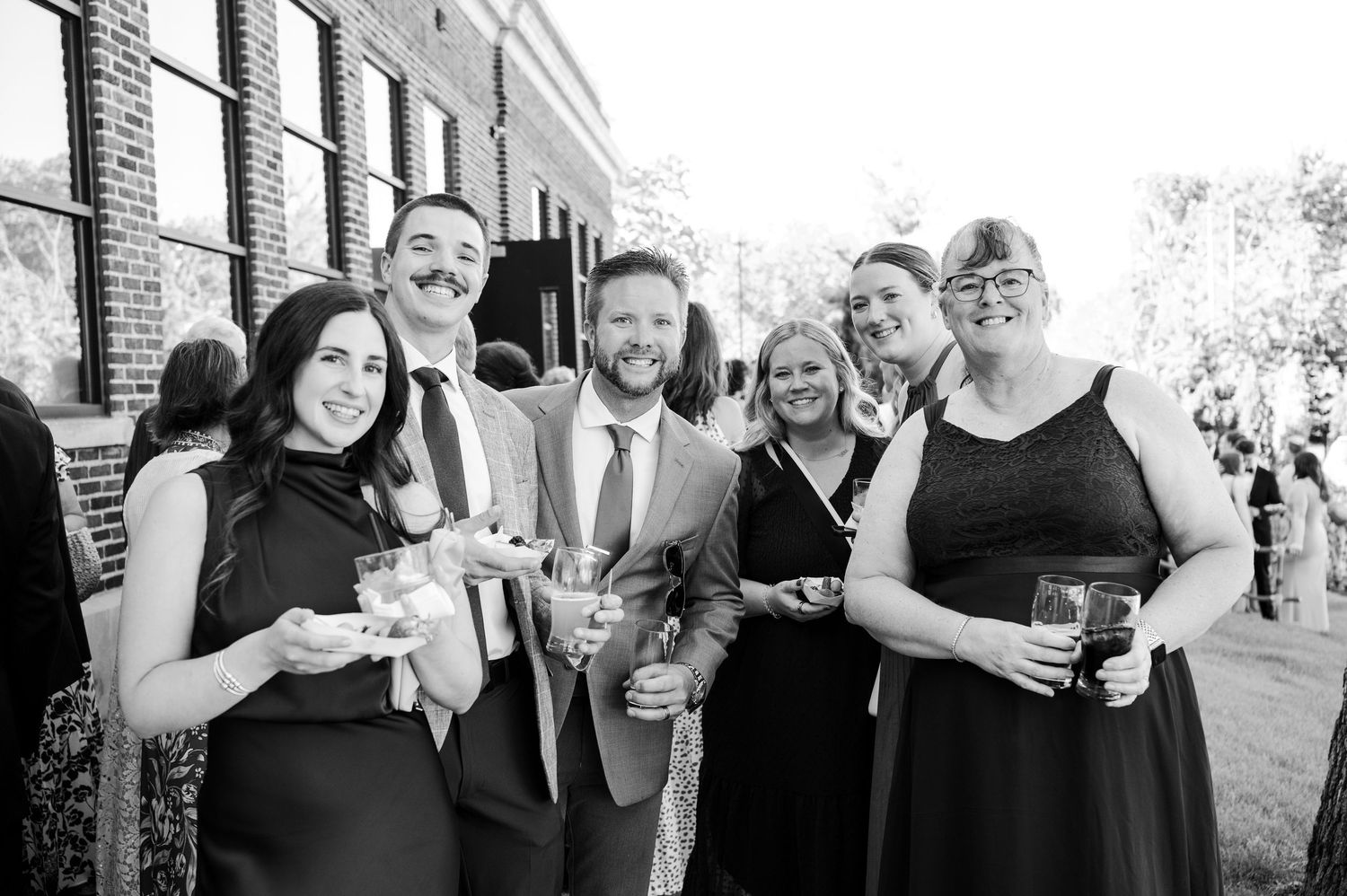 Black and white photo of five smiling friends dressed in formal attire holding drinks at an outdoor wedding reception.