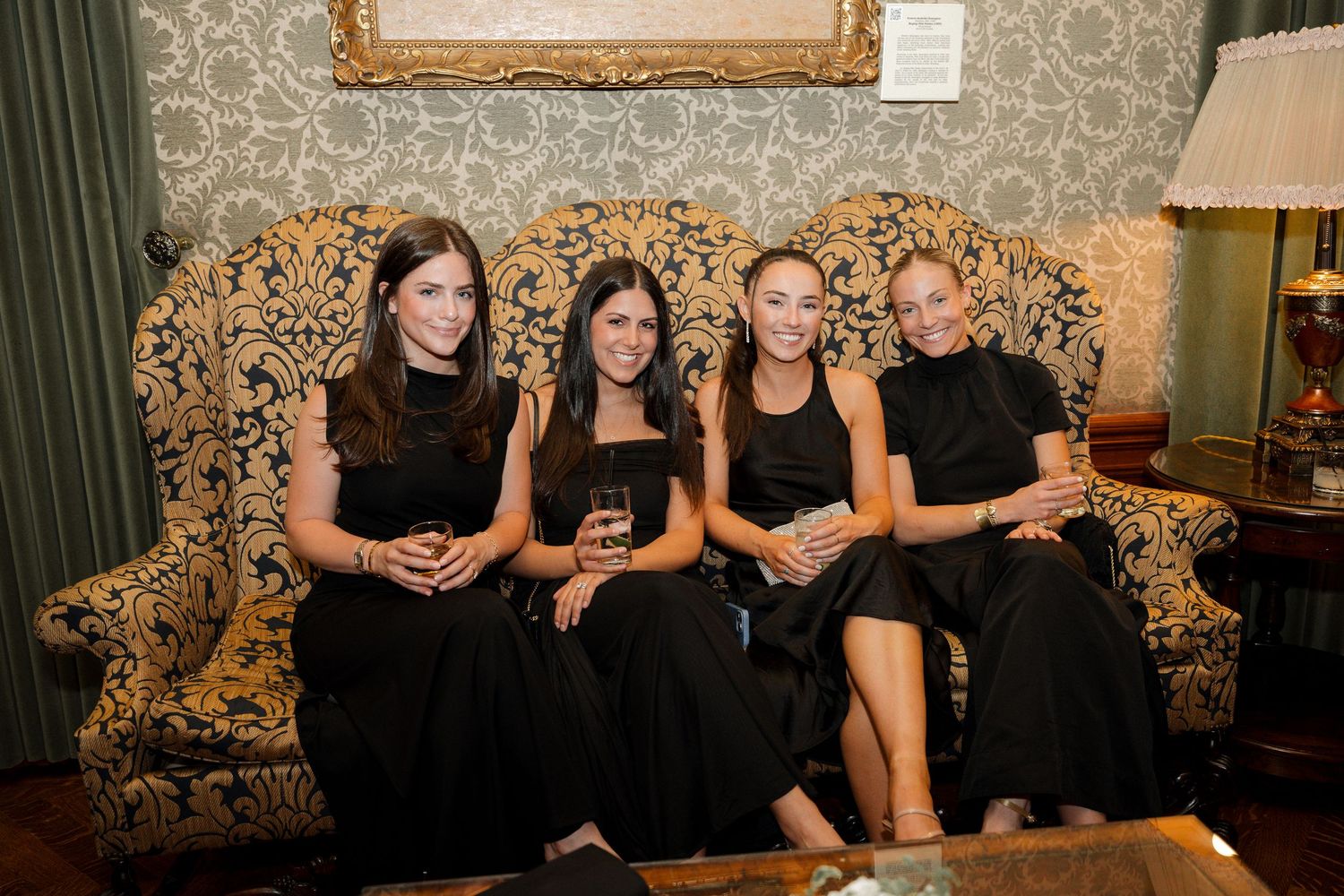 Four women in black dresses sitting together on an ornate sofa at an elegant wedding reception.
