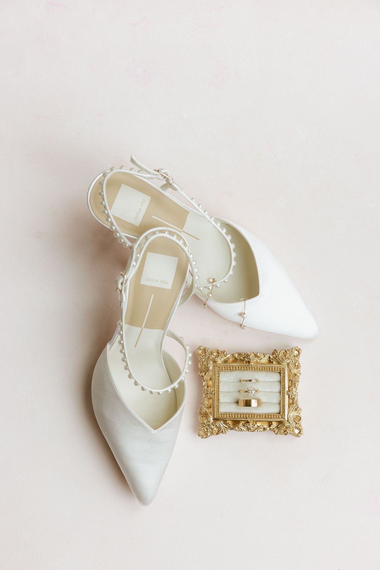 White pearl-strap bridal heels displayed with gold ornate frame holding wedding rings.