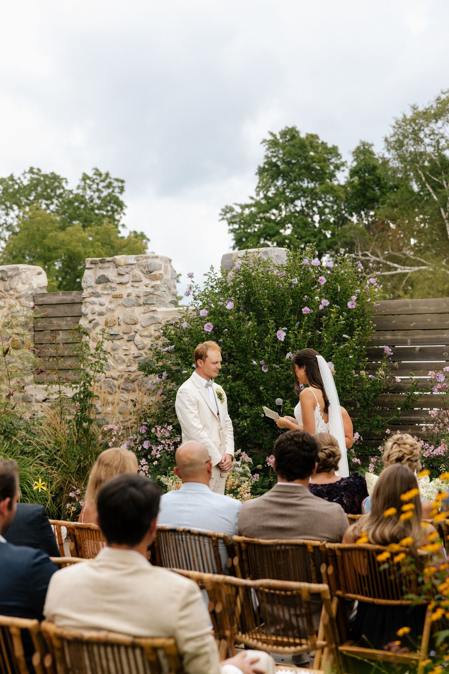 Outdoor wedding ceremony with bride and groom at rustic stone wall altar, guests seated in wooden chairs amid lush garden.