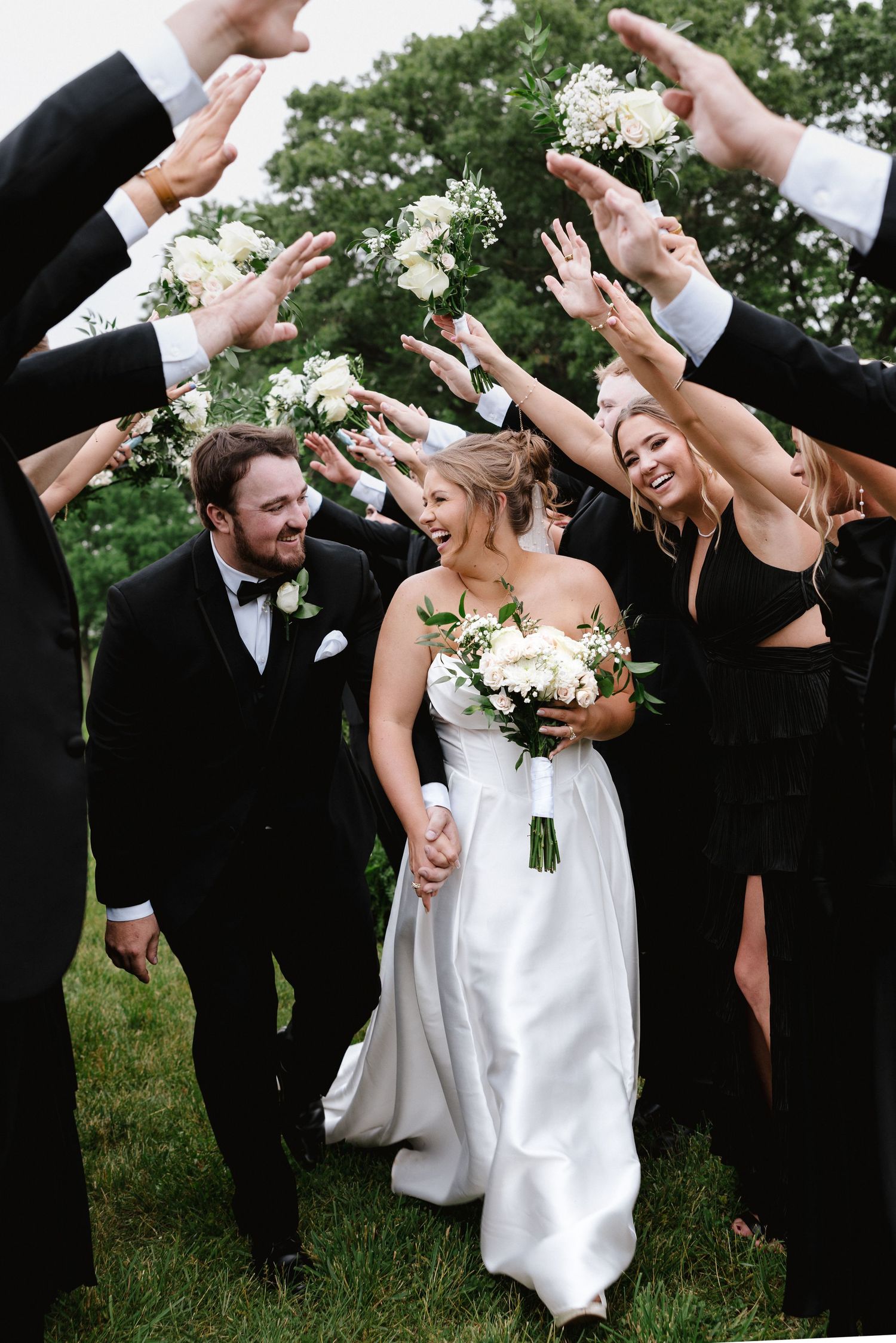 Bride and groom walk through arch of raised bouquets held by wedding party in black attire outdoors.