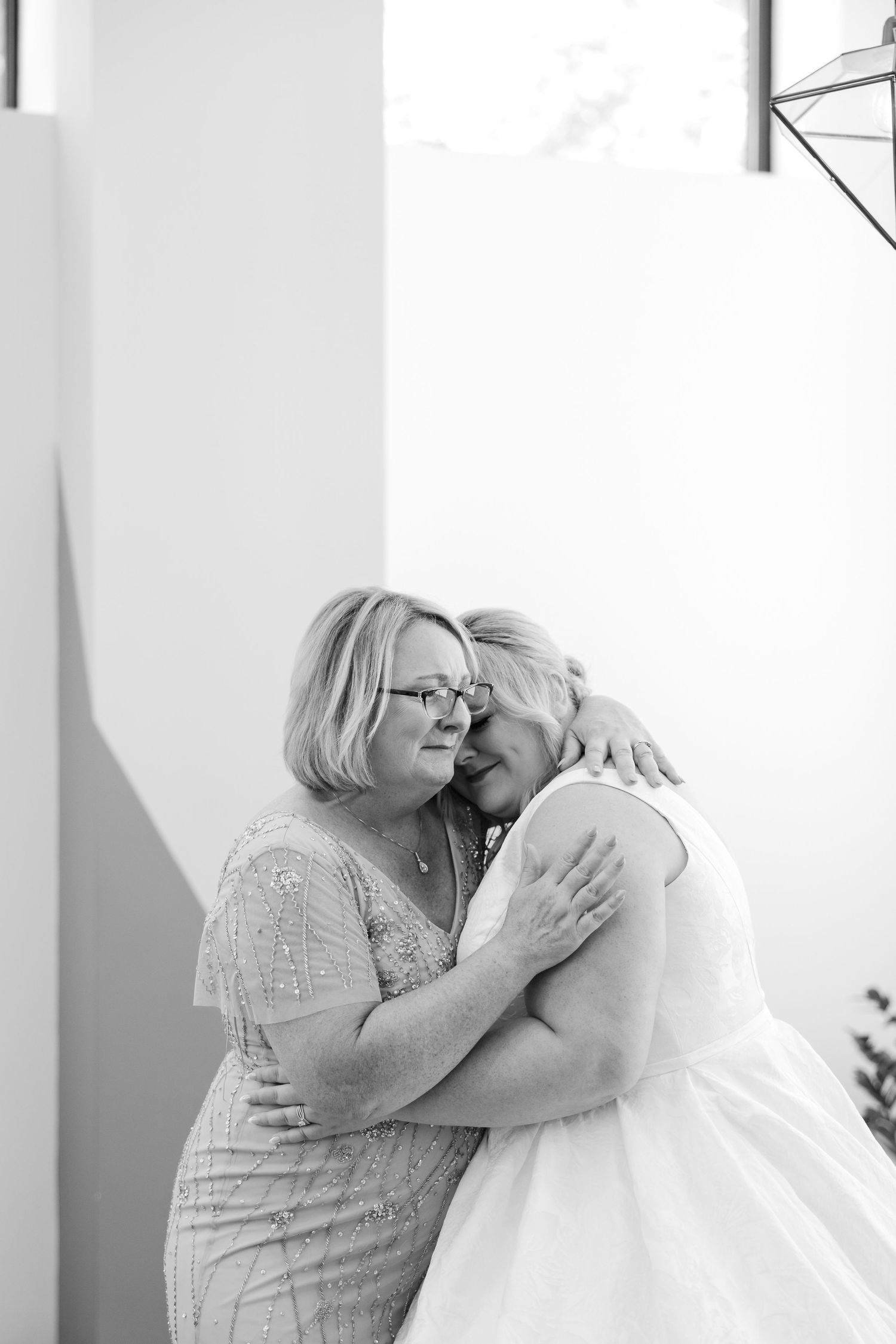 Black and white photo of a bride in white dress sharing an emotional hug with an older woman in a beaded gown.