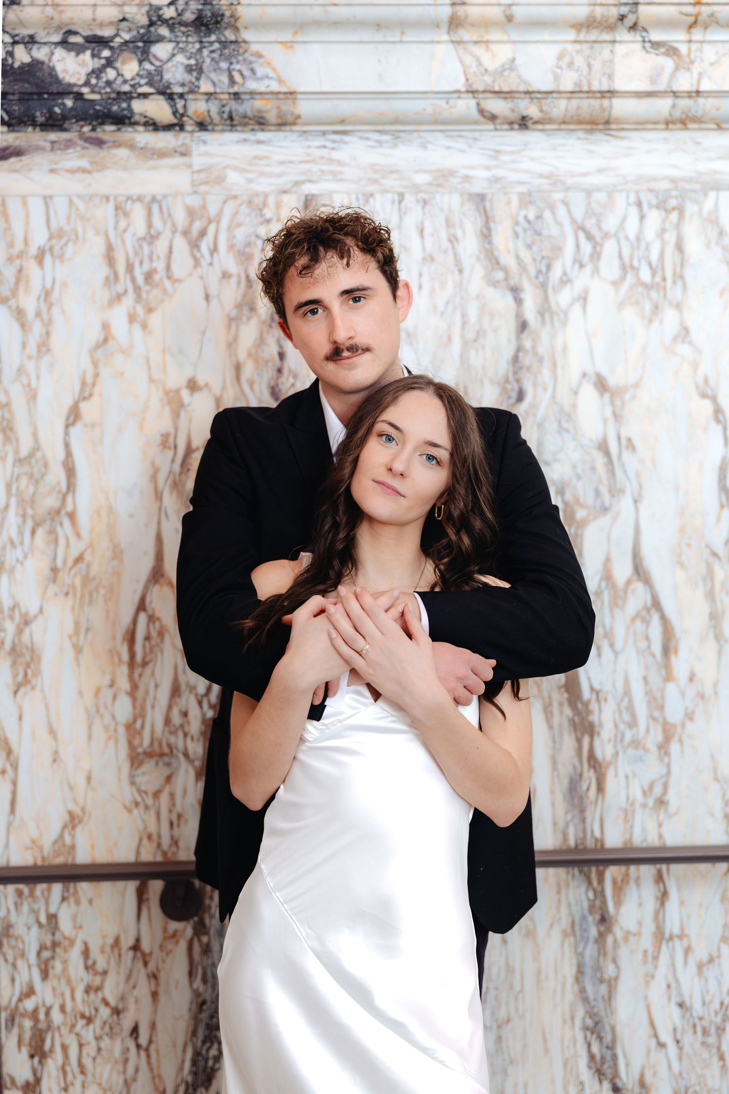 Bride in white satin dress and groom in black suit embracing before marble columns in elegant wedding portrait.