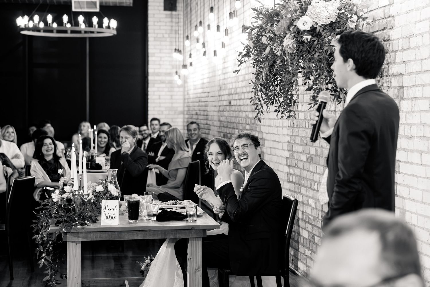 Black and white photo of wedding guests laughing during a groom speech at a candlelit reception table.