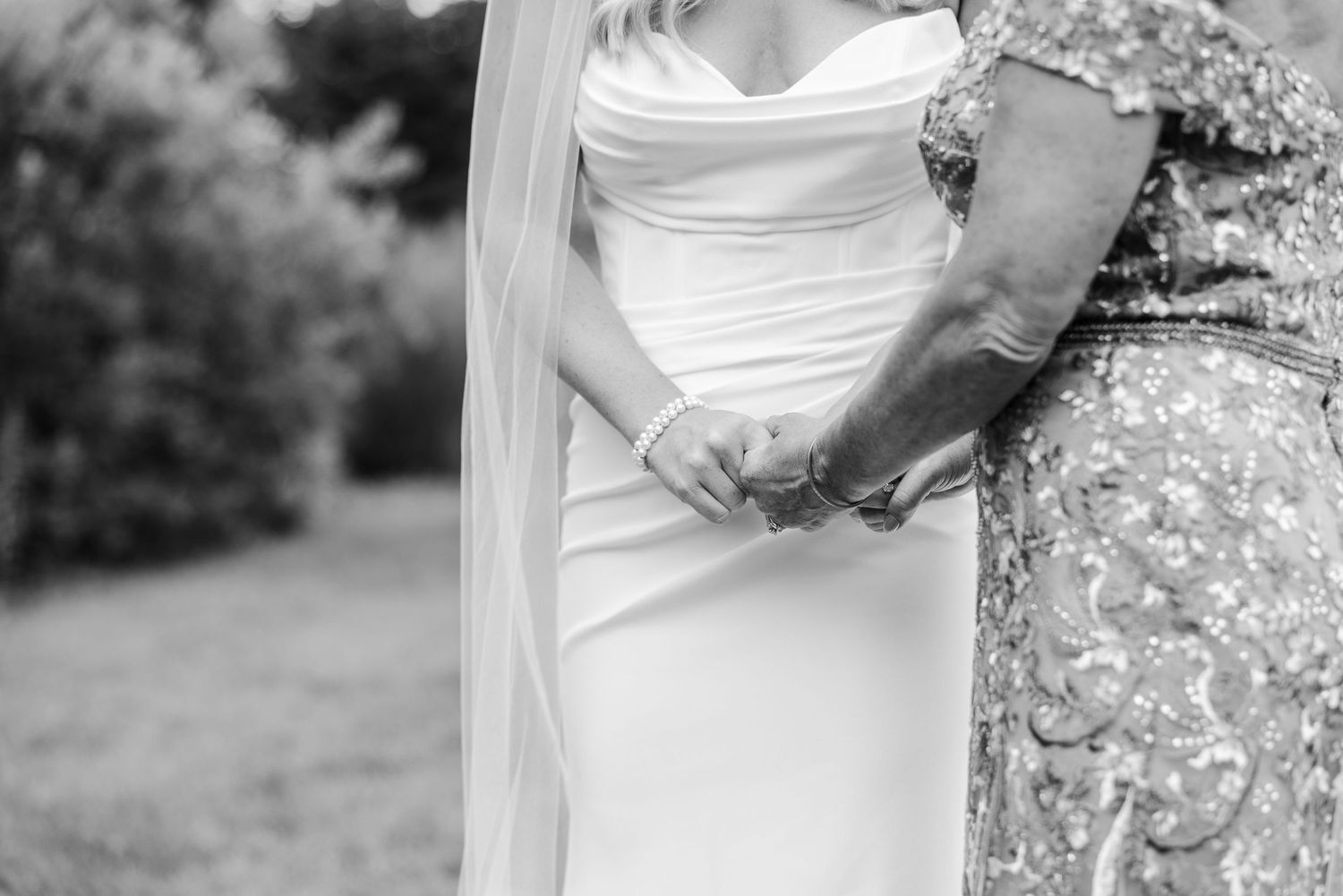 Black and white photo of a bride in a white gown and veil holding hands with an older woman in a beaded dress outdoors.