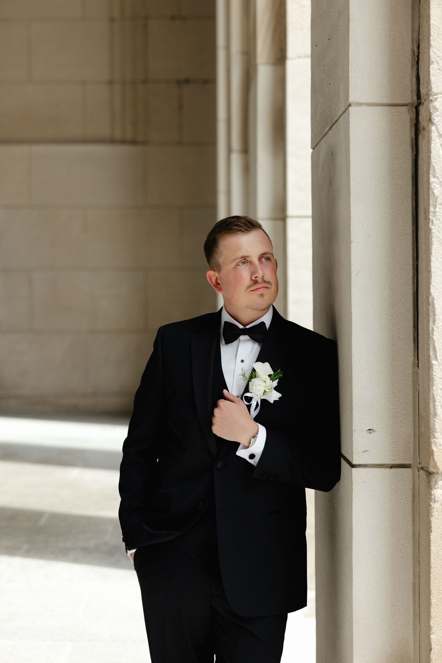 A dapper groom in a black tuxedo with white boutonniere leans against a stone column, gazing upward.