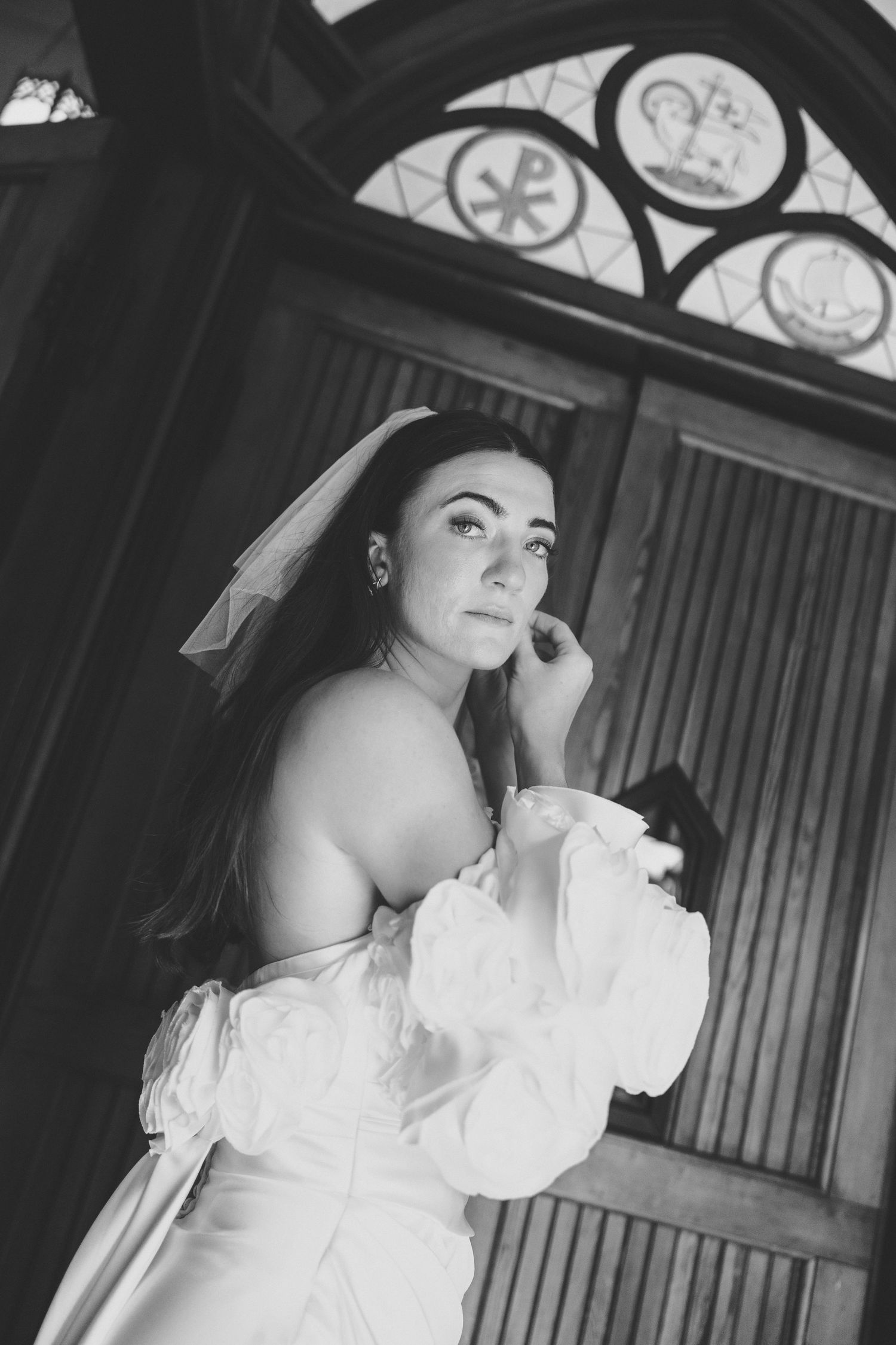 Black and white portrait of a bride in a white gown and veil standing before ornate church doors.