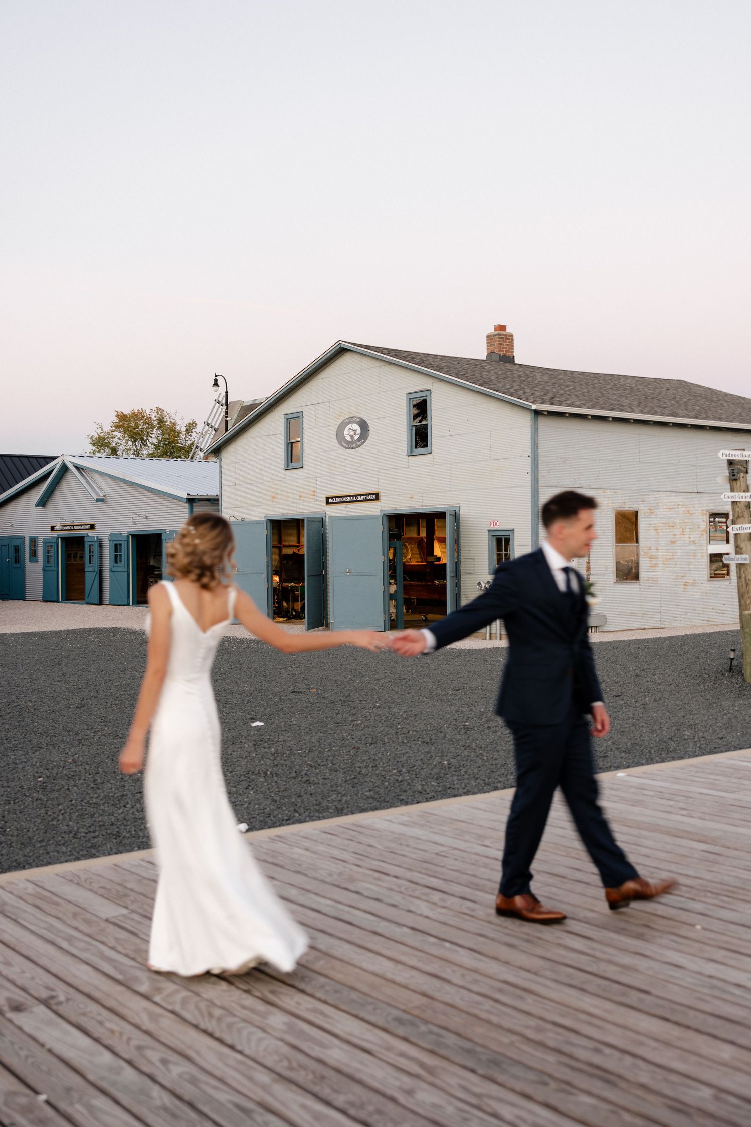 Bride and groom hold hands walking on wooden boardwalk at dusk, white barn venue in background.