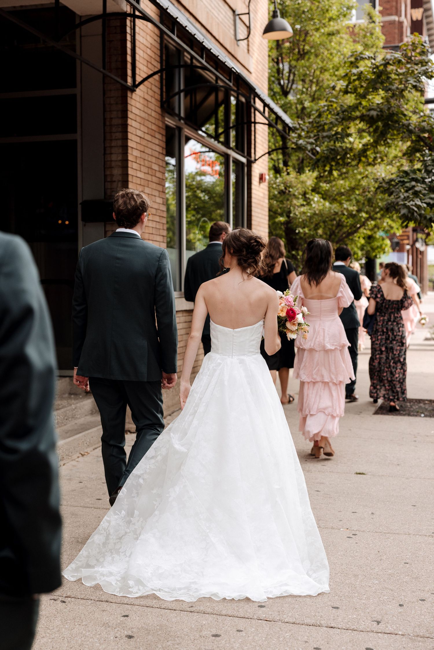 Bride in strapless ball gown walks with groom and guests along urban sidewalk.