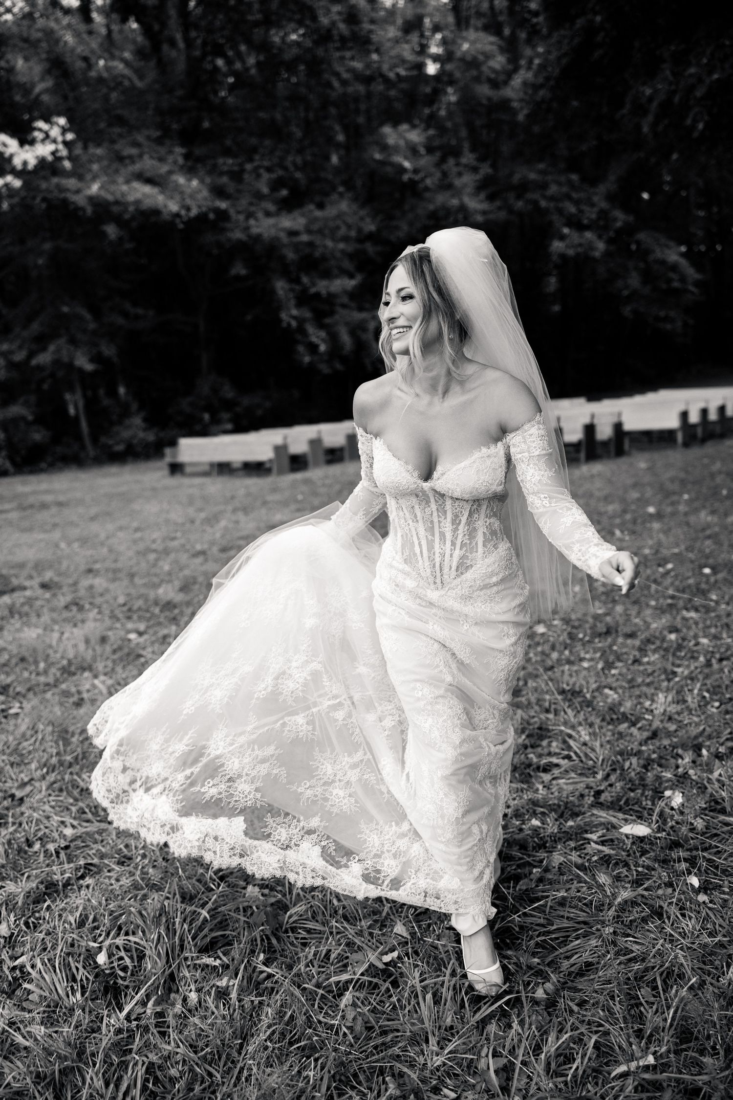 Black and white photo of a smiling bride in a lace wedding gown and veil running across a grassy lawn.