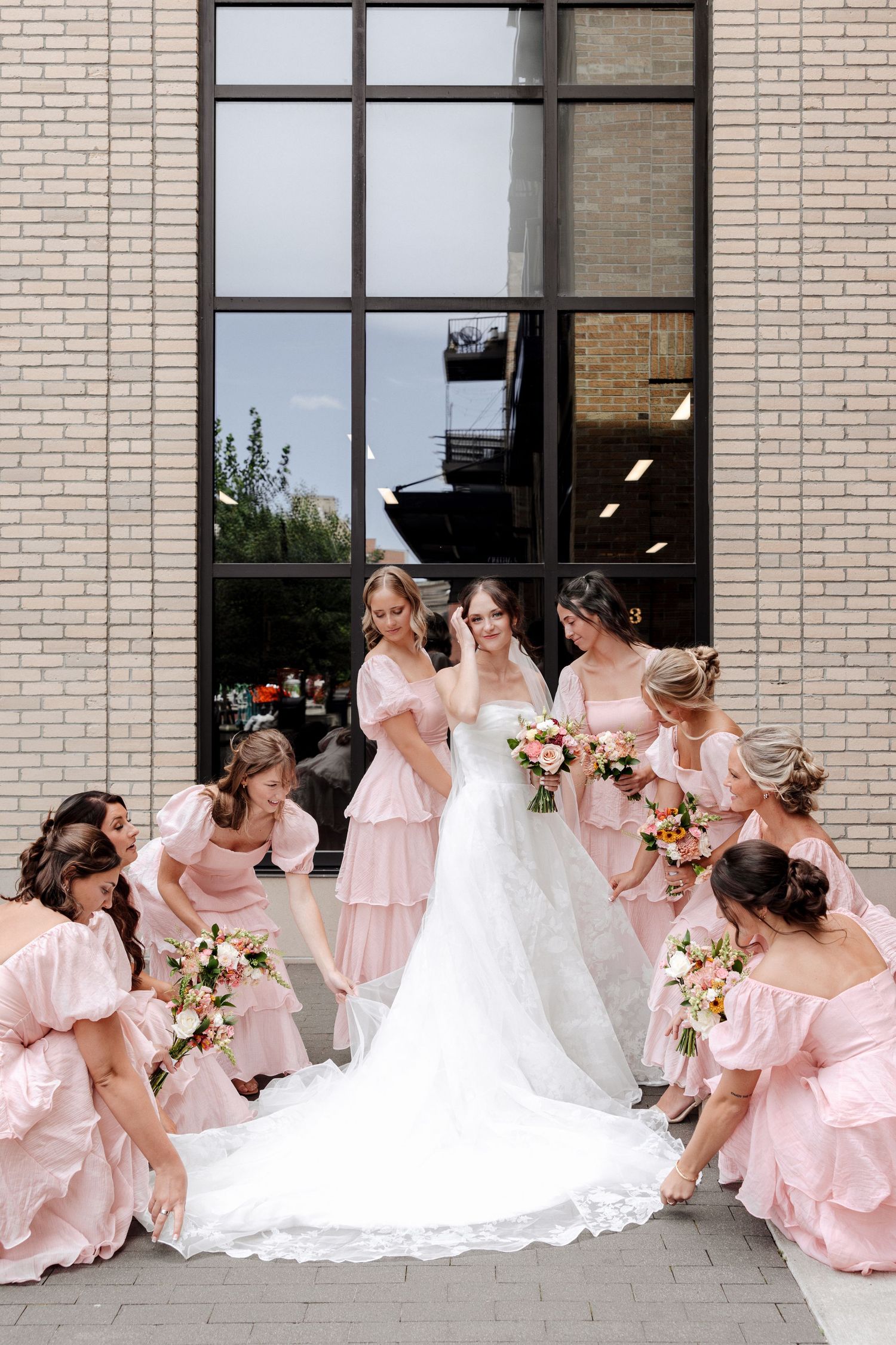 Bride in white lace gown surrounded by bridesmaids in pink dresses arranging her train outside brick building.