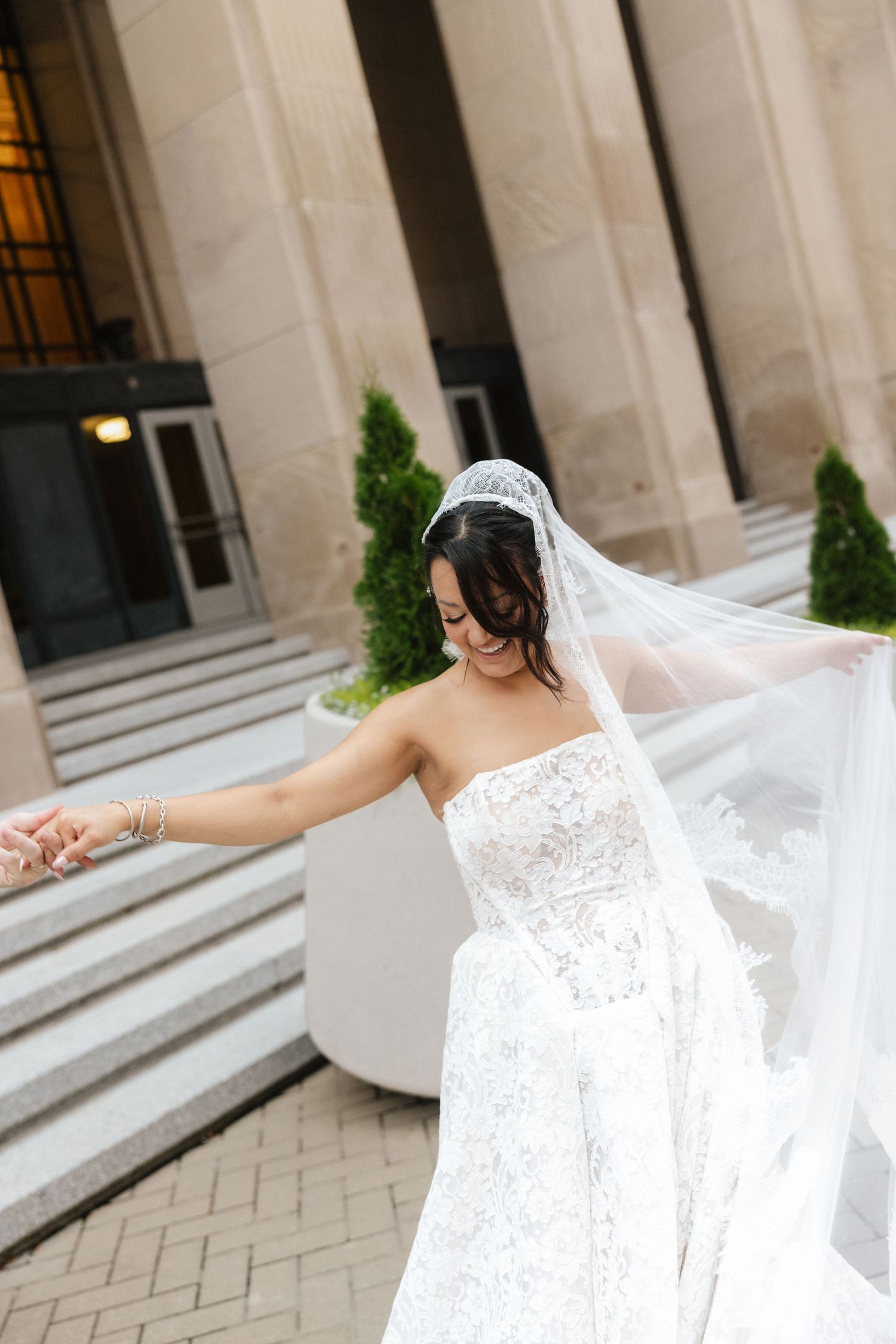 Bride in strapless lace wedding gown with flowing veil twirling outside elegant building with marble steps.