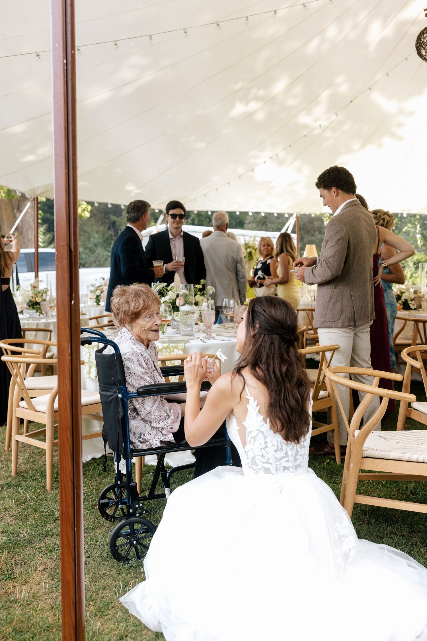 Bride in white gown kneeling to speak with elderly guest in wheelchair at outdoor wedding.
