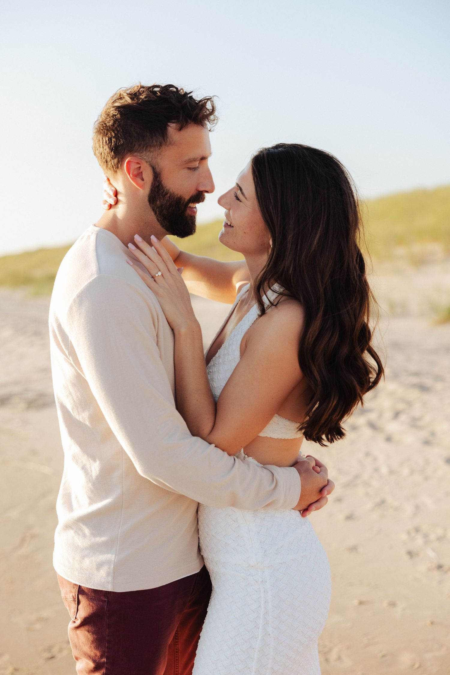 A couple embraces romantically on a sandy beach, the woman in a white dress and man in a beige shirt gazing into each other's eyes.