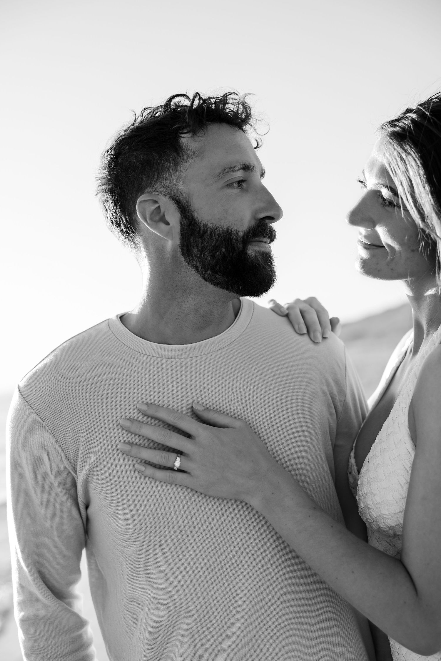 Black and white photo of a bearded man and smiling woman embracing outdoors, she rests hand on his chest.