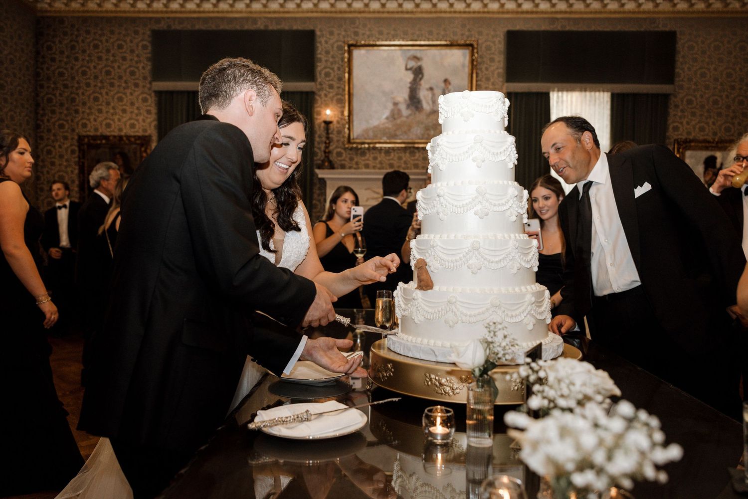 Wedding guests in black tie attire gather around a tall white tiered wedding cake during reception.