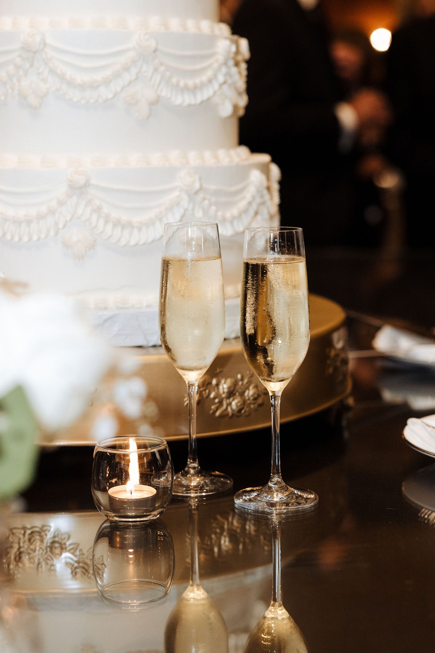 Two champagne flutes beside a white multi-tiered decorated wedding cake with a lit candle.