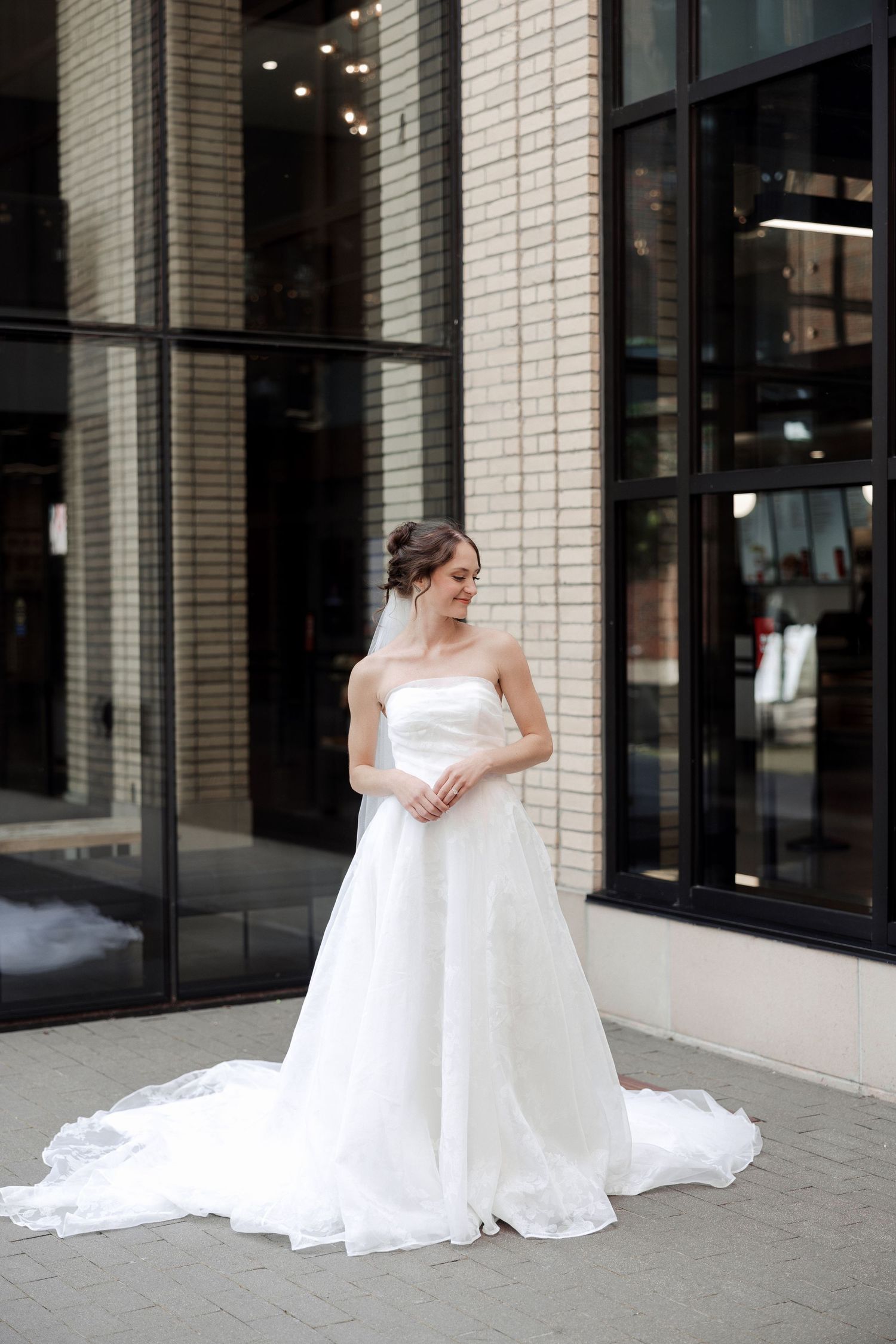Elegant bride in a strapless white ball gown with a long train stands outside a modern brick building.