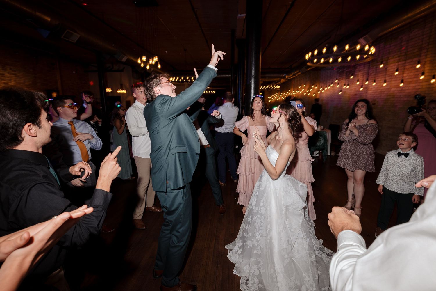 Bride and groom dance joyfully surrounded by guests at a lively indoor wedding reception.