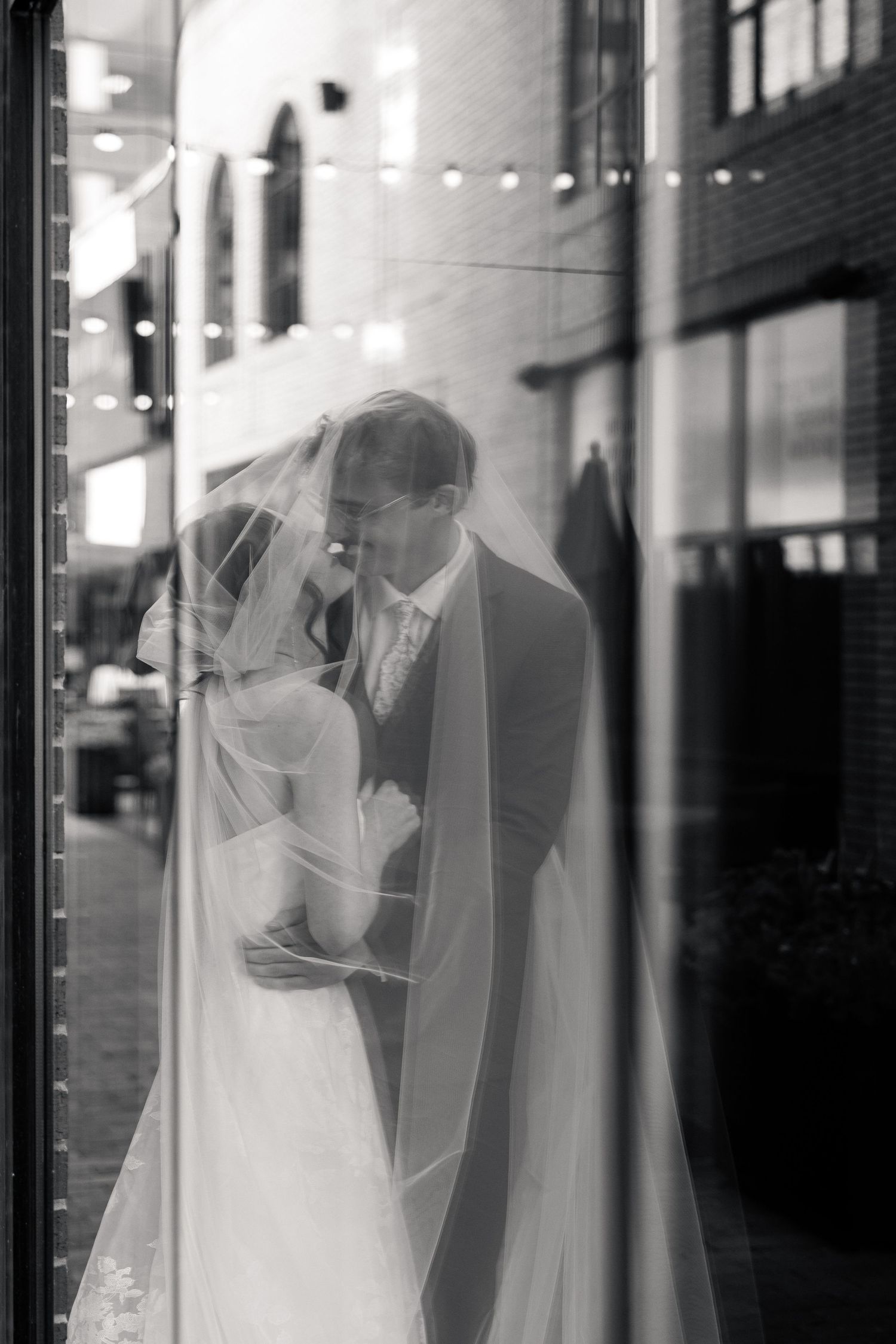Romantic black and white reflection of a bride and groom embracing beneath a veil in a glass window.
