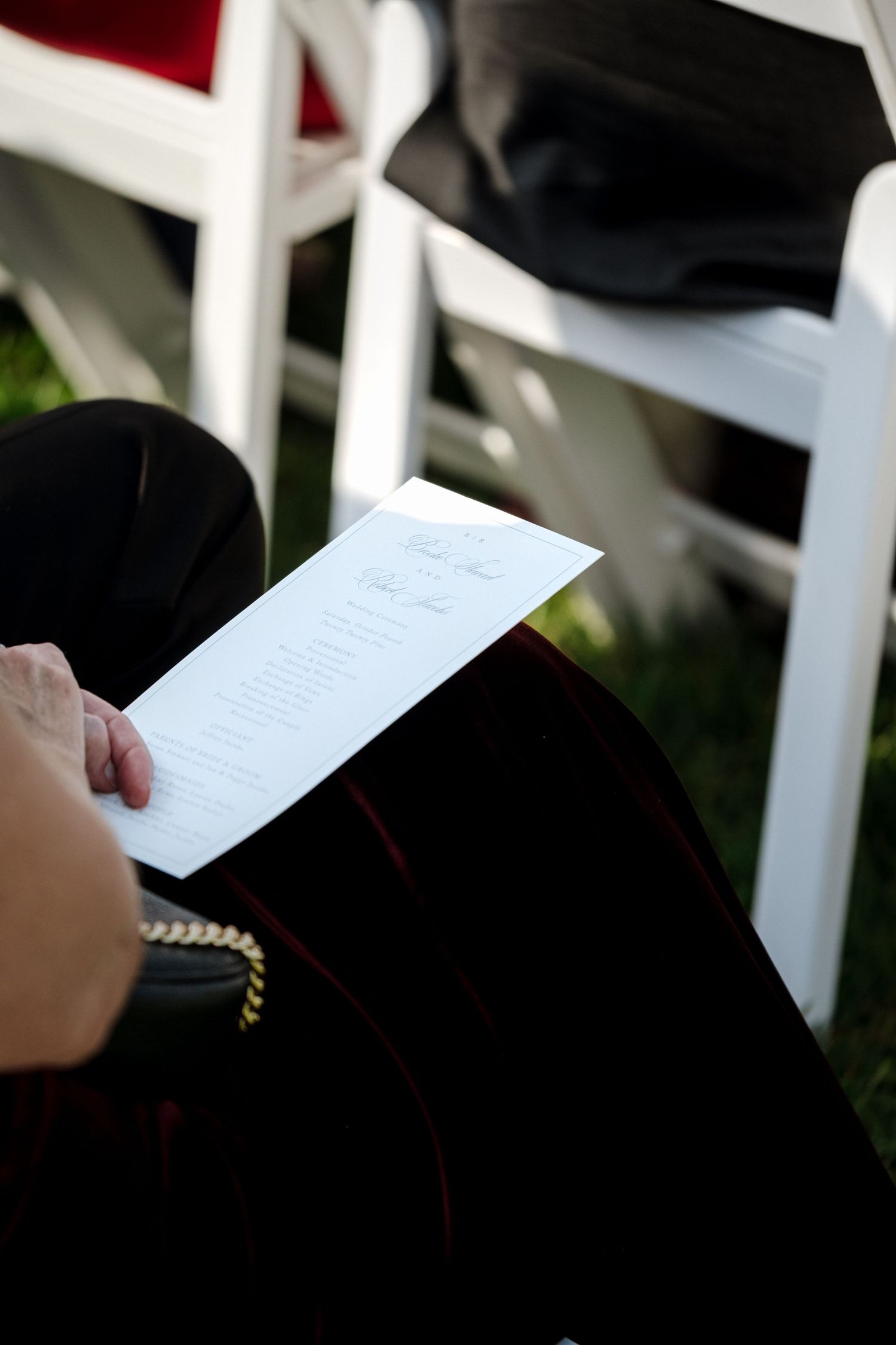 Wedding guest reading ceremony program while seated in white chair at outdoor wedding ceremony.