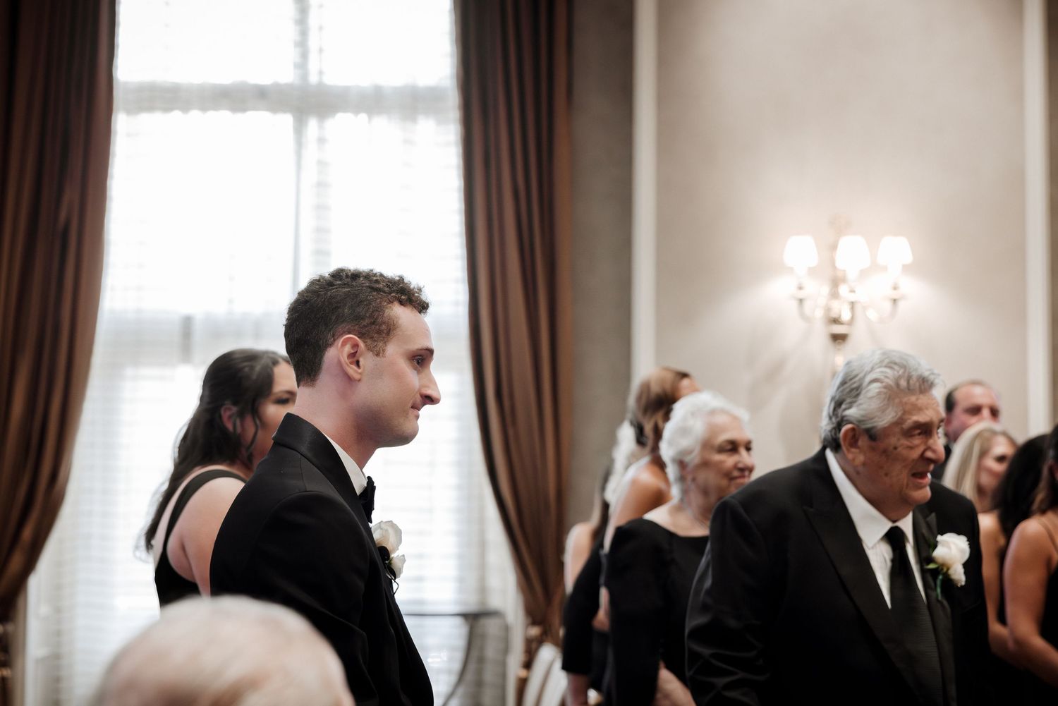 Groom in black tuxedo looks emotional as he watches his bride walk down the aisle during the wedding ceremony.