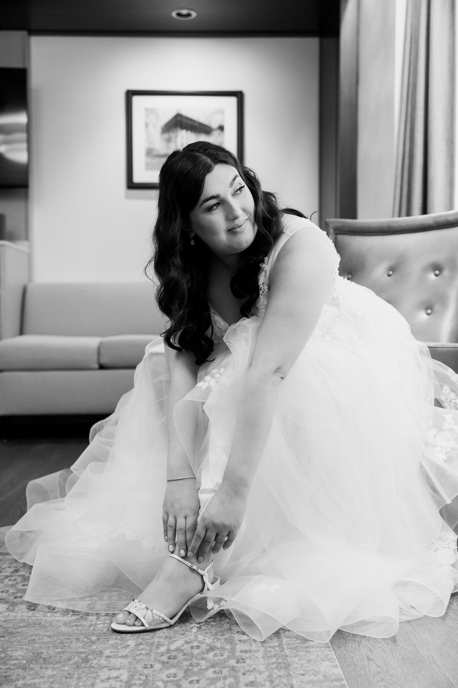 Black and white photo of bride in white gown putting on heels while seated on a tufted chair.