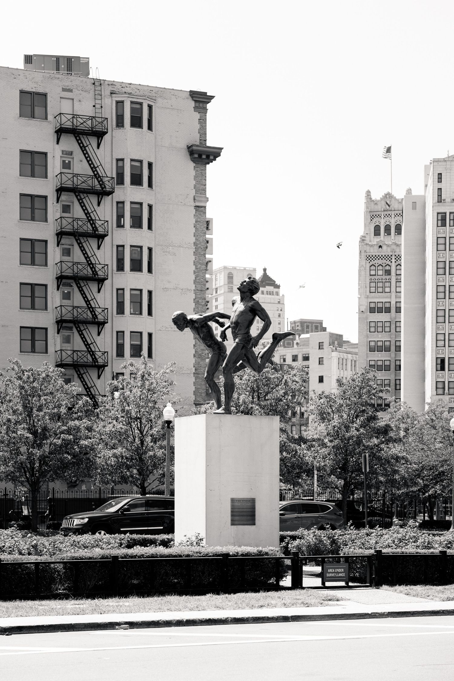 Black and white photo of a bronze sculpture on a pedestal along a city boulevard with tall buildings.